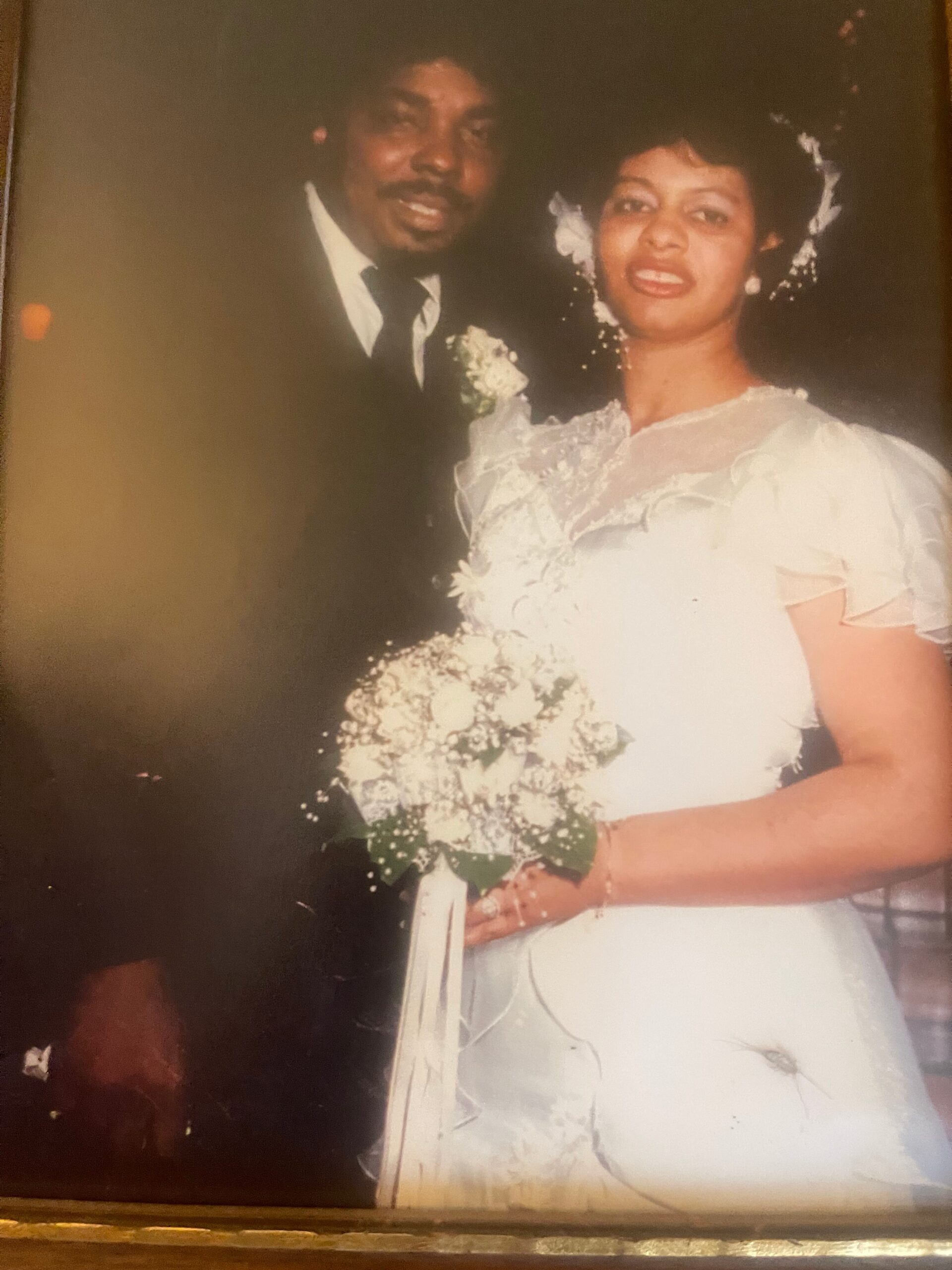Bride in a lace wedding dress holds a bouquet of white flowers, standing with a groom in a dark suit and tie.