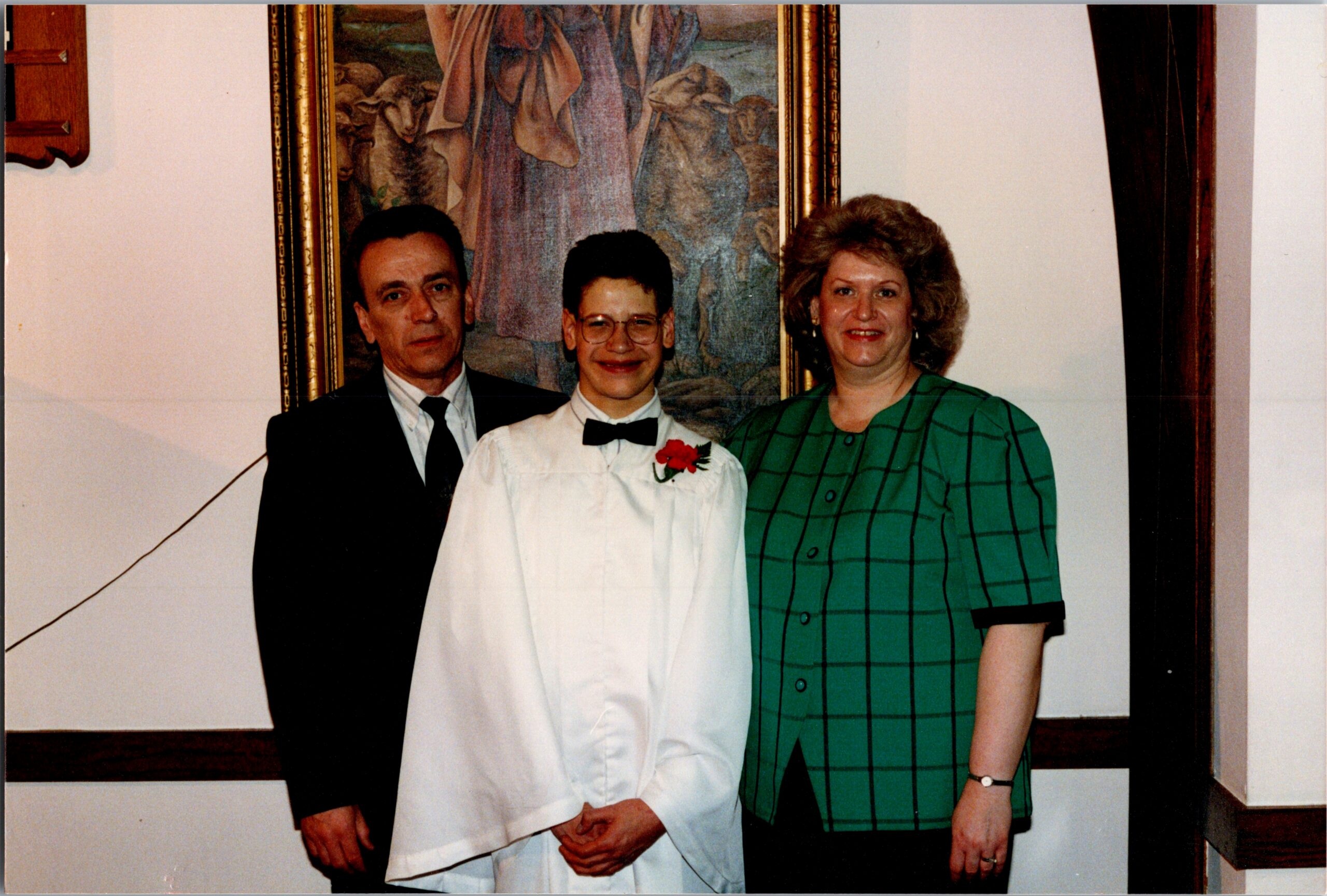 Three people posing indoors: an older man in a suit on the left, a young man in a white shirt with bow tie and red carnation in the center, and a woman in a green plaid dress on the right, with a framed painting behind them.