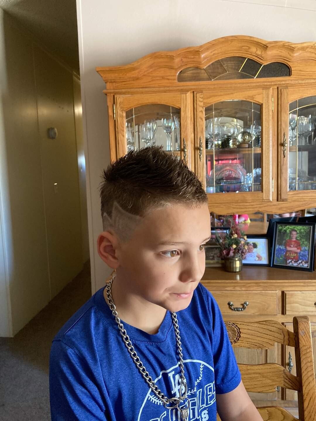 Boy with a short, spiked haircut and shaved sides wearing a blue shirt and thick chain necklace, seated in a dining room with a wooden china cabinet behind him.