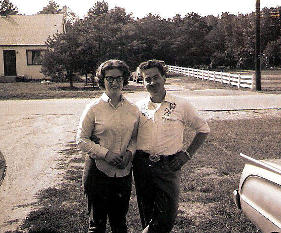 Two young adults pose together in a rural driveway, arms linked, with a house and trees in the background.