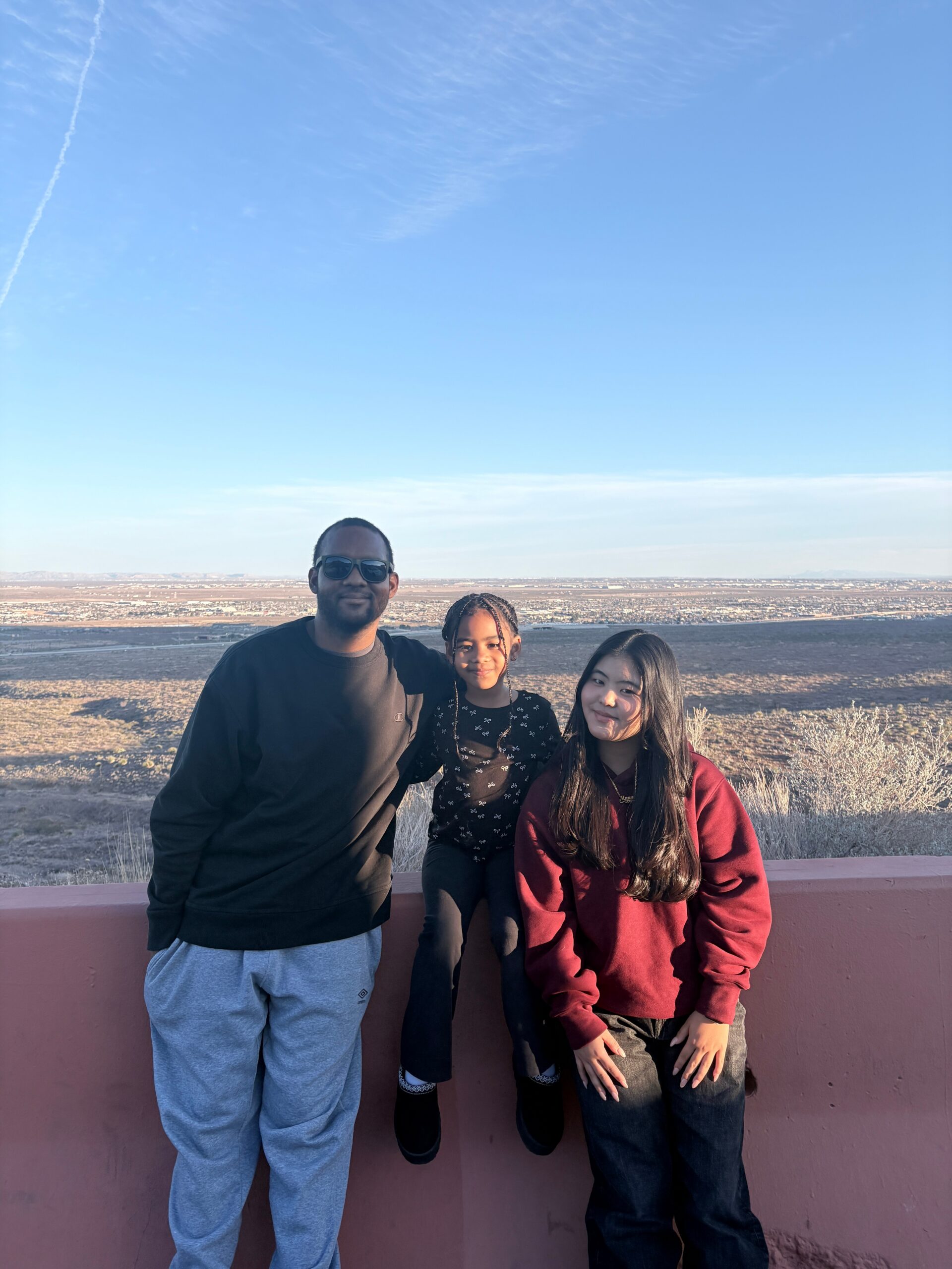 Three people posing together outdoors in front of an arid desert landscape under a clear blue sky, smiling at the camera.