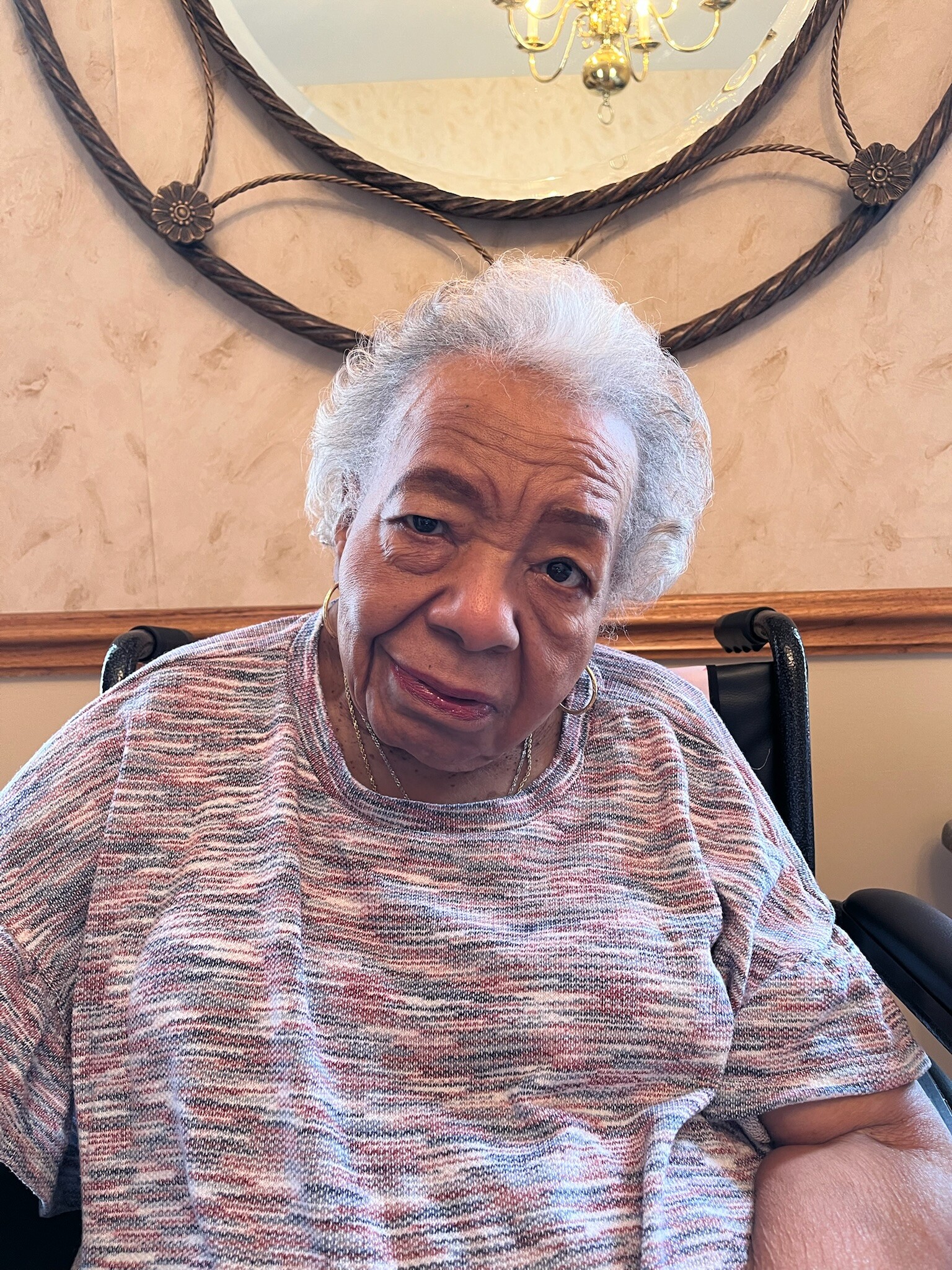 Elderly woman with short gray hair, wearing a pink-gray striped sweater, seated indoors in a wheelchair with a decorative mirror and chandelier behind her.