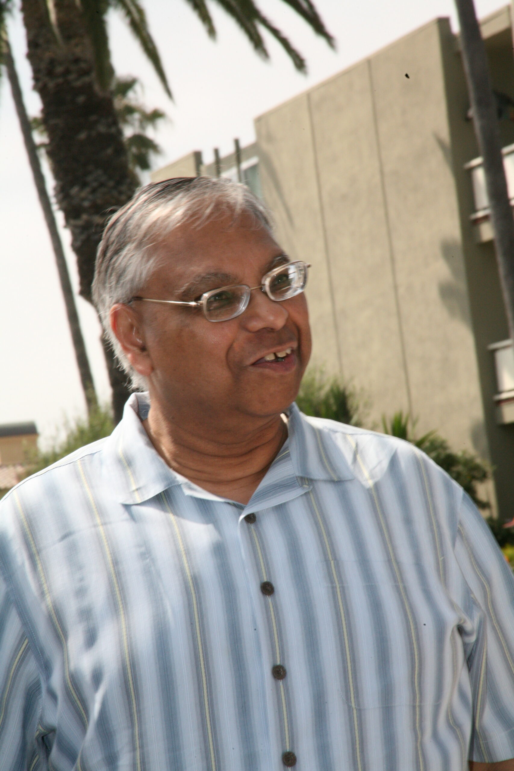 Smiling man with gray hair and glasses, wearing a light blue striped shirt, outdoors with palm trees in the background.