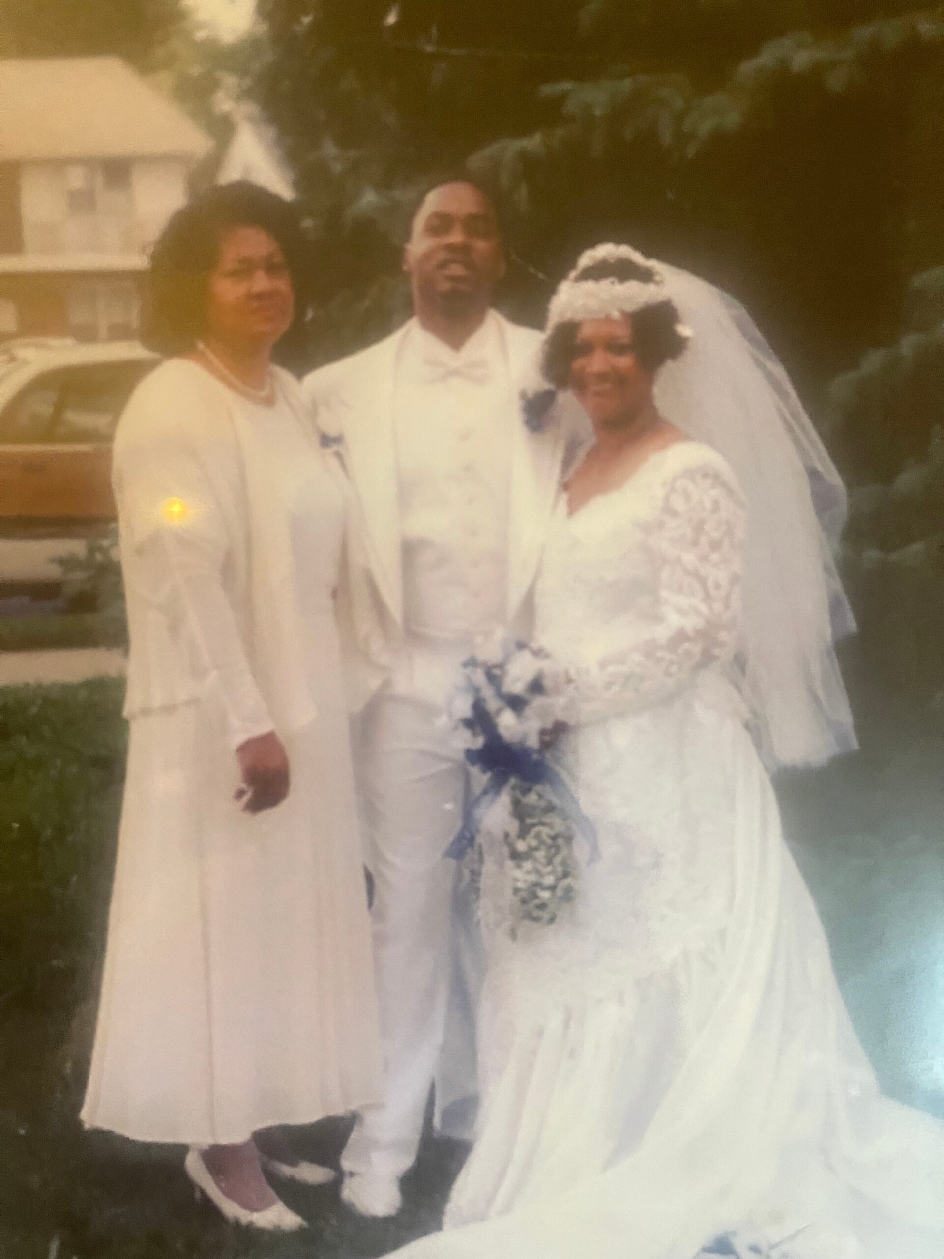 Bride in a lace wedding dress and veil stands with groom in a white suit and bow tie, flanked by a guest in white, outdoors with greenery behind them.
