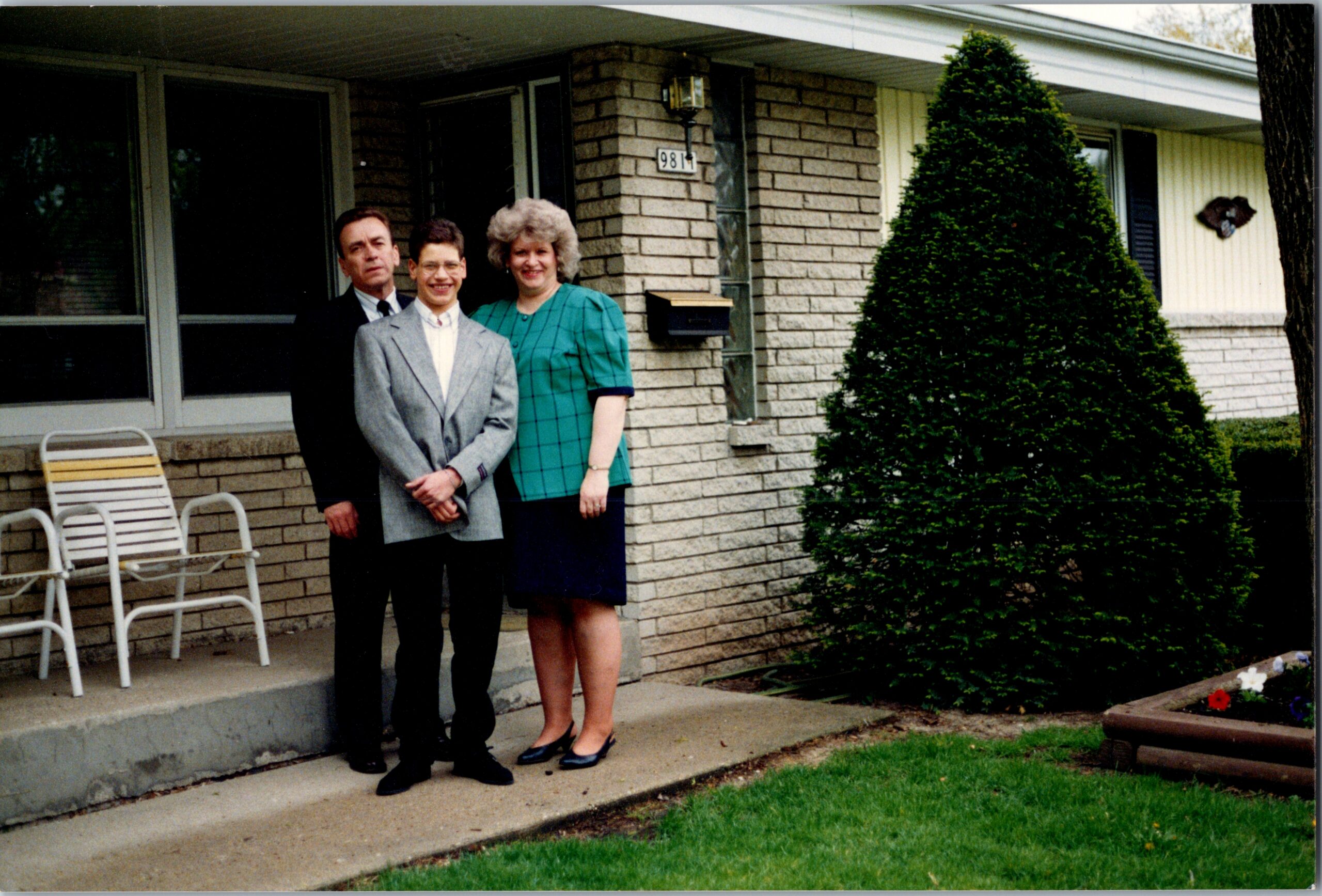 Three adults pose on a brick house porch; a man in a gray suit, a man in a dark suit, and a woman in a teal dress smile at the camera (house number 9811 visible). he