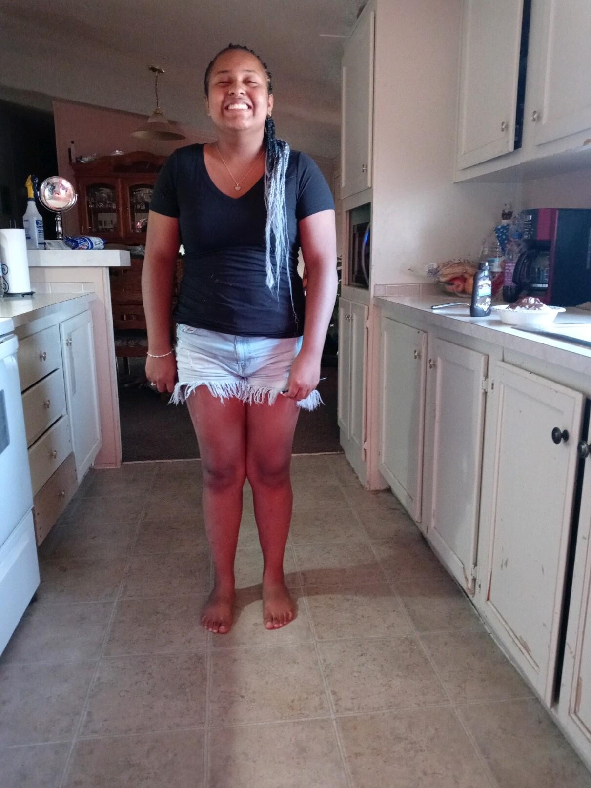 Woman standing barefoot in a kitchen, smiling with eyes closed, wearing a black t-shirt and frayed denim shorts.