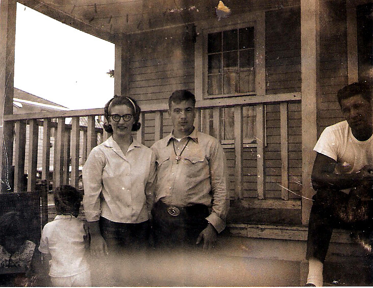 Three adults pose on a weathered wooden porch; a woman with glasses stands beside a man in a button-down shirt, with another man in a white T-shirt at the right.