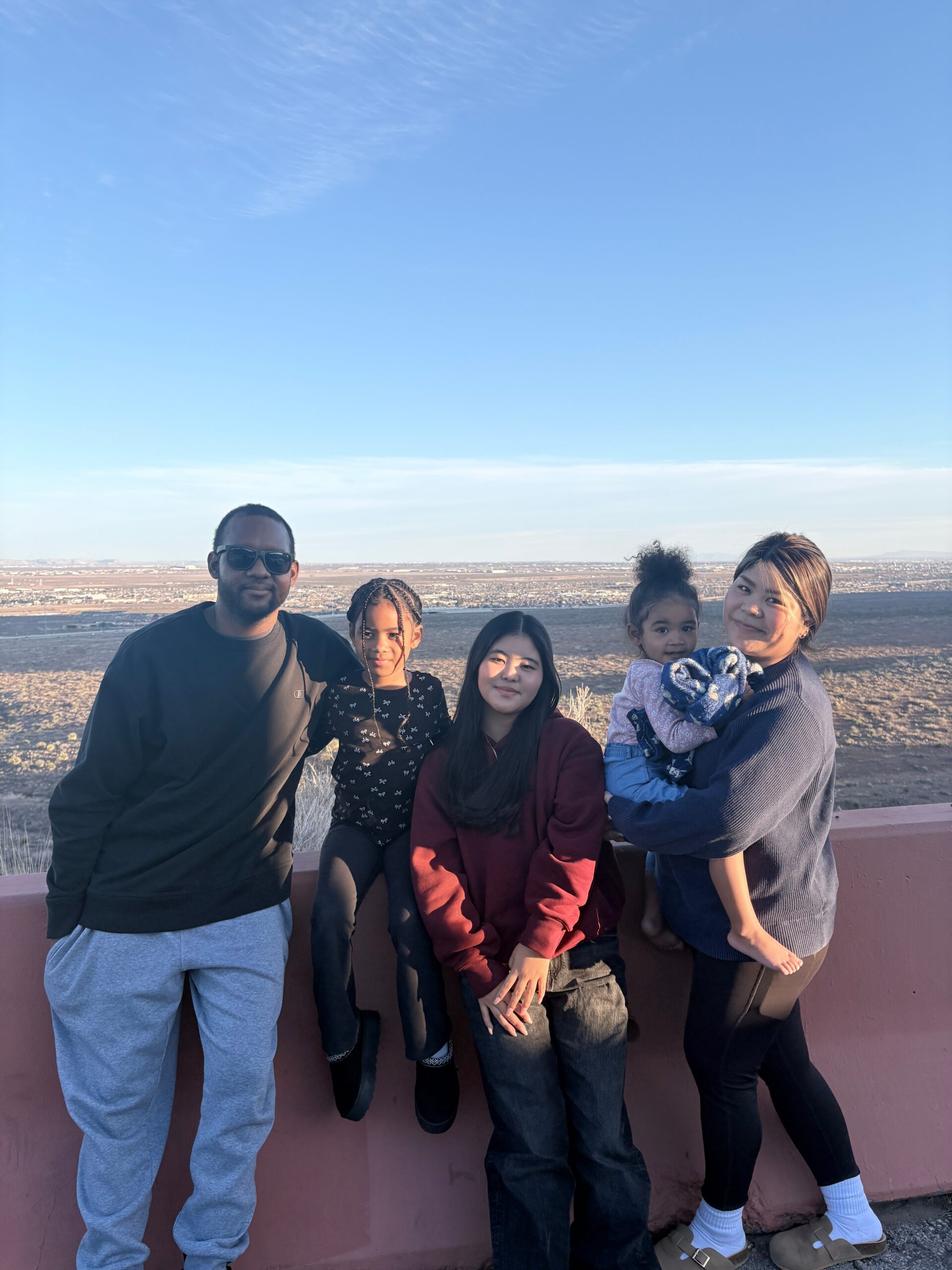 Family group of six posing outdoors on a sunny day with a desert landscape in the background.