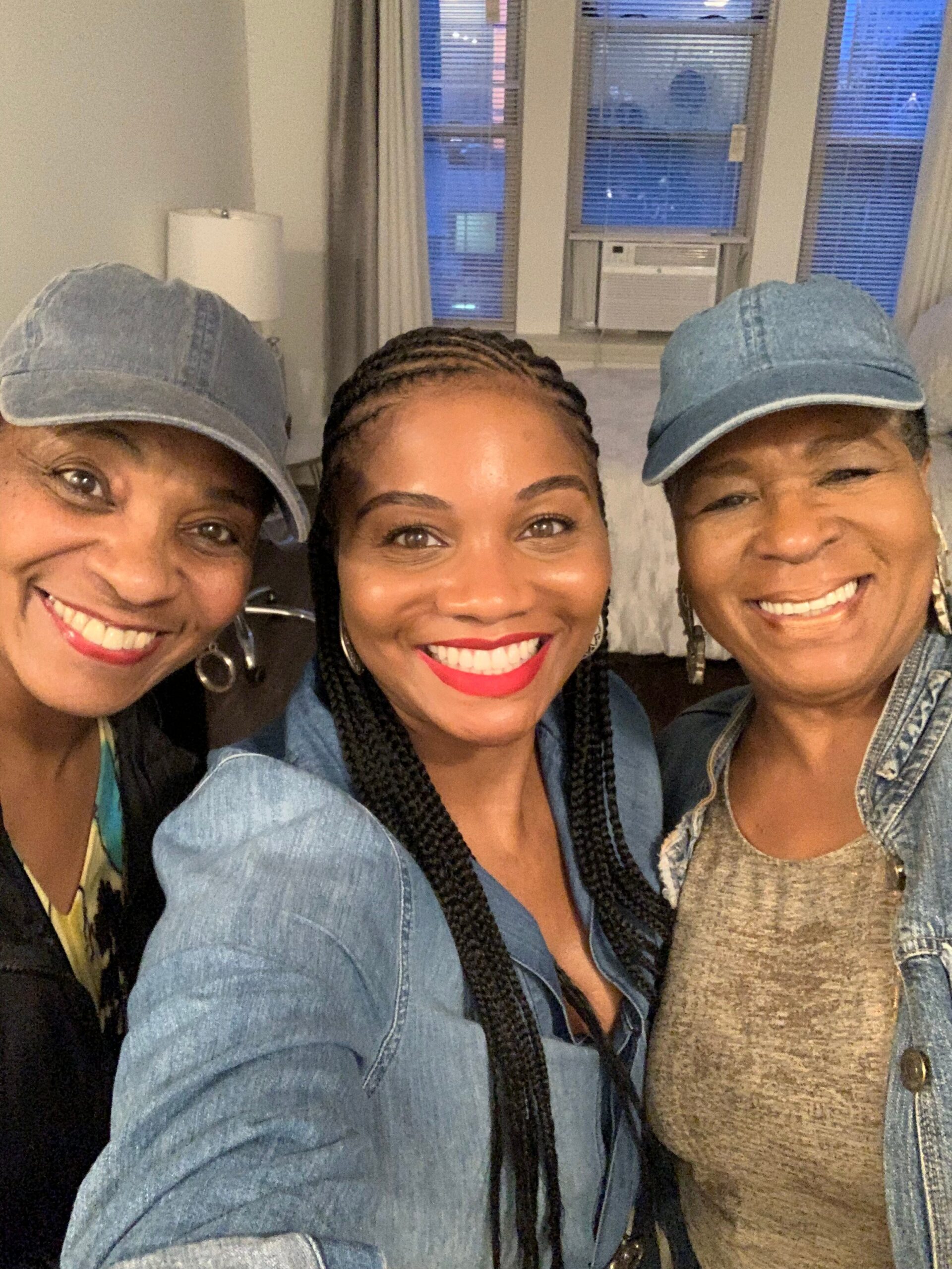 Three smiling women take a selfie indoors, wearing denim jackets and blue caps. A window AC unit and blinds appear in the background.