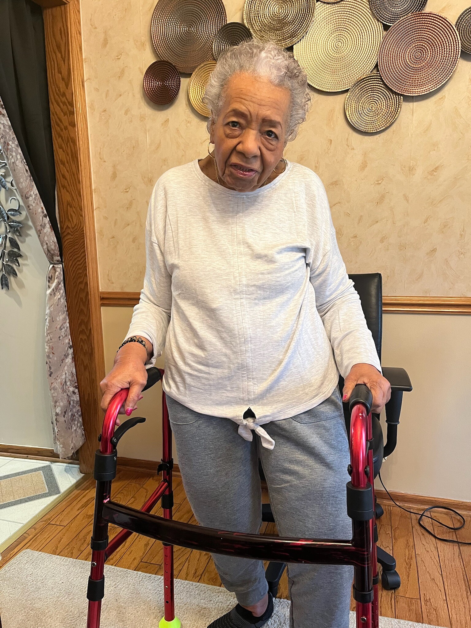 Elderly woman standing behind a red mobility walker indoors, looking at the camera.