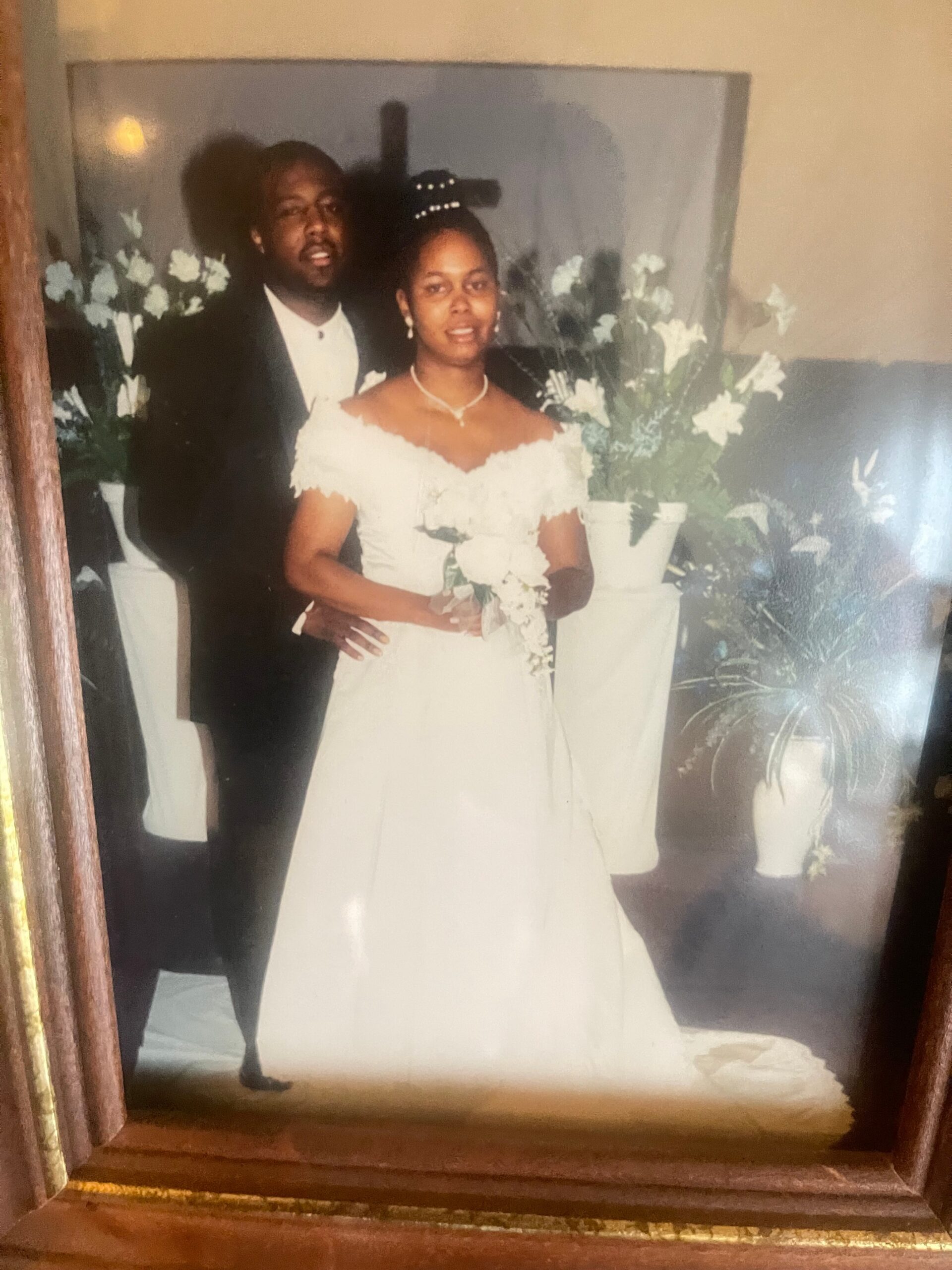 Bride in an off-shoulder white wedding gown holding a bouquet, with a groom in a suit standing behind her amid white florals.