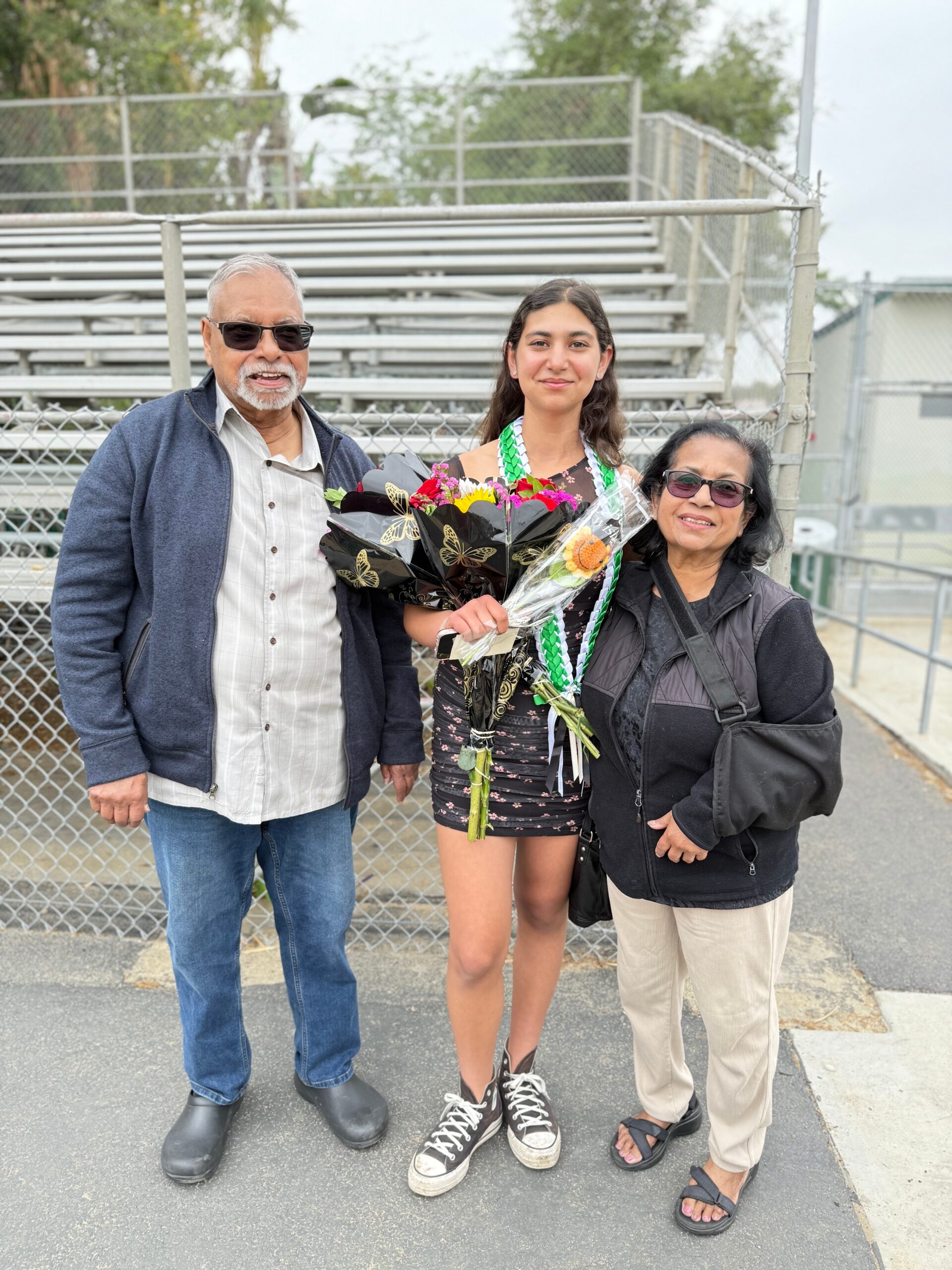 Three people posing for a photo outdoors by bleachers: an elderly man in a blue jacket and sunglasses, a young woman with flowers and a green lei, and an older woman in black with glasses.
