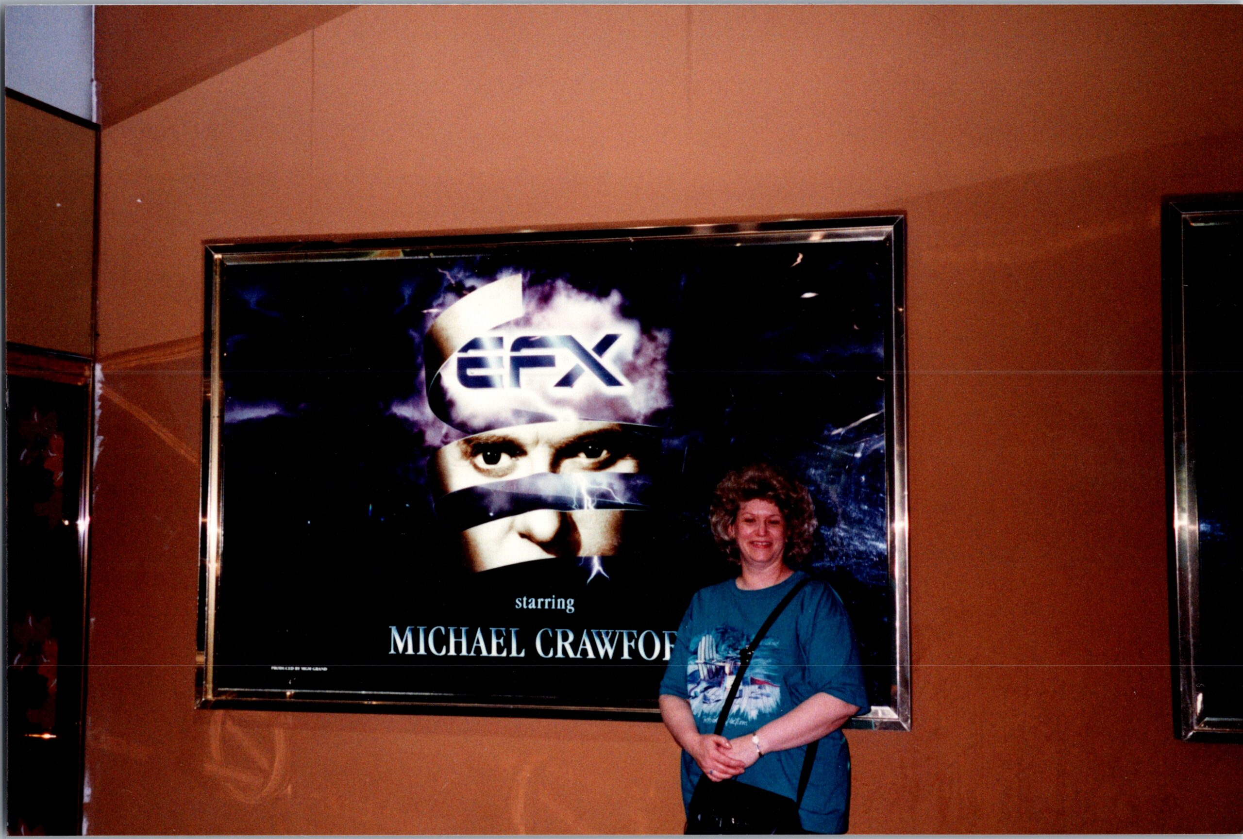 Woman in a teal shirt standing in front of a framed movie poster on an orange wall.