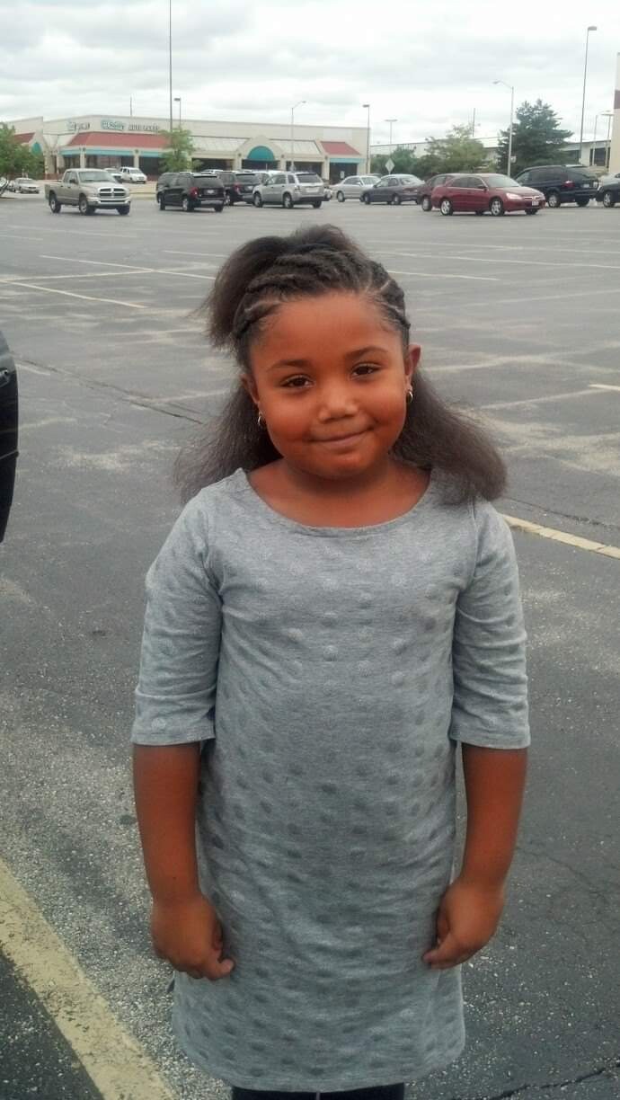 Young girl with braided hair wearing a gray knee-length dress stands in a parking lot, with a shopping plaza in the background.