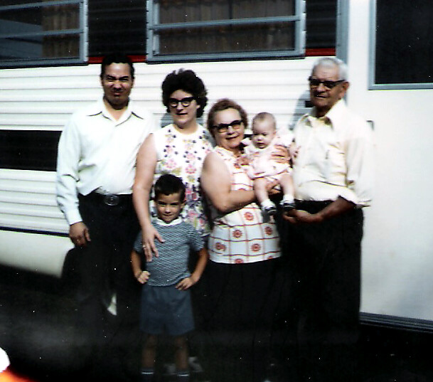 A multi-generational family group posing in front of a mobile home, including two children and two adults holding a baby.