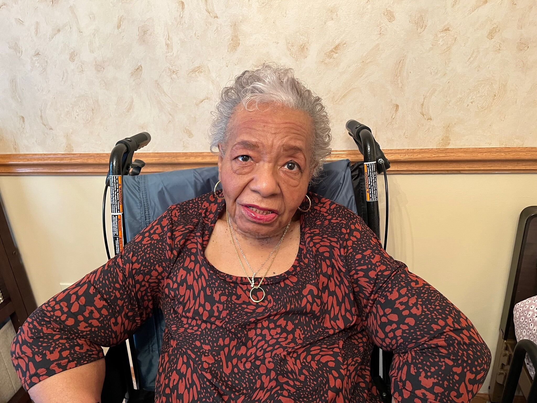 Older woman in a wheelchair smiling at the camera, wearing a red and black patterned blouse indoors.