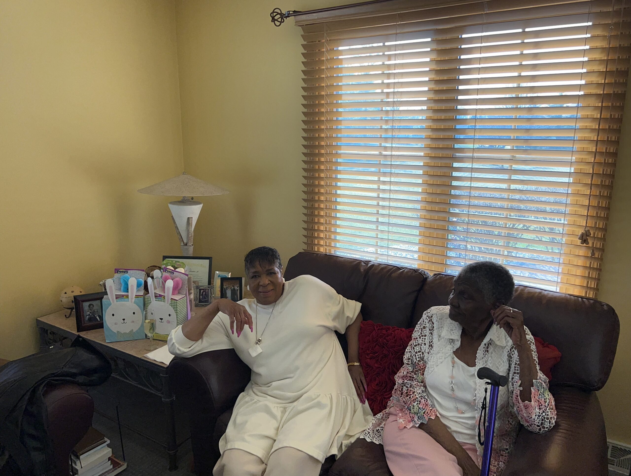 Two elderly women sit on a brown leather sofa in a living room, smiling, with bunny-themed gift bags and photos on a side table nearby.