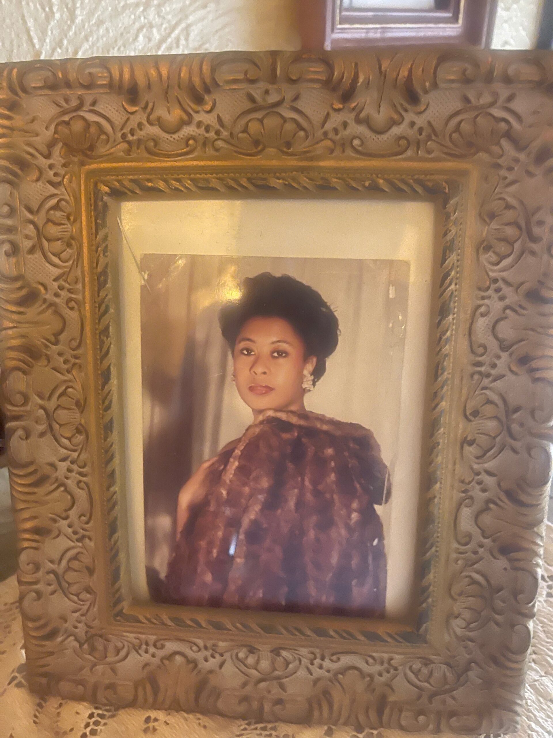 Formal portrait of a woman in a dark, patterned dress, framed by an ornate gold frame with floral motifs in a vintage setting.