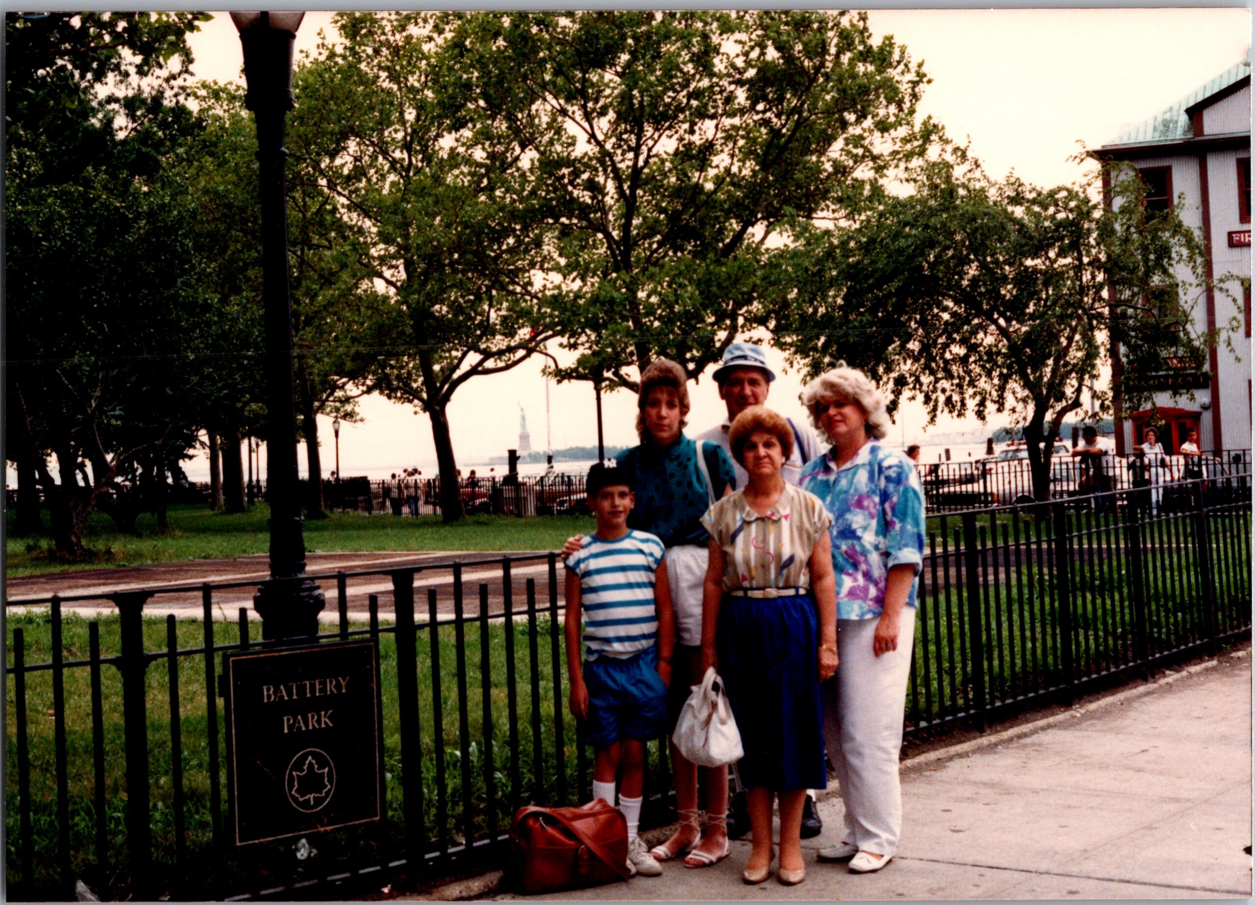 Group of people posing in front of a black iron fence at Battery Park, with trees and a waterfront promenade in the background.