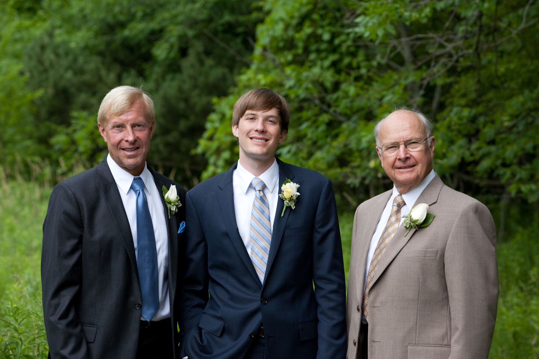Three men in formal suits posing outdoors among green trees; boutonnieres visible on their lapels.