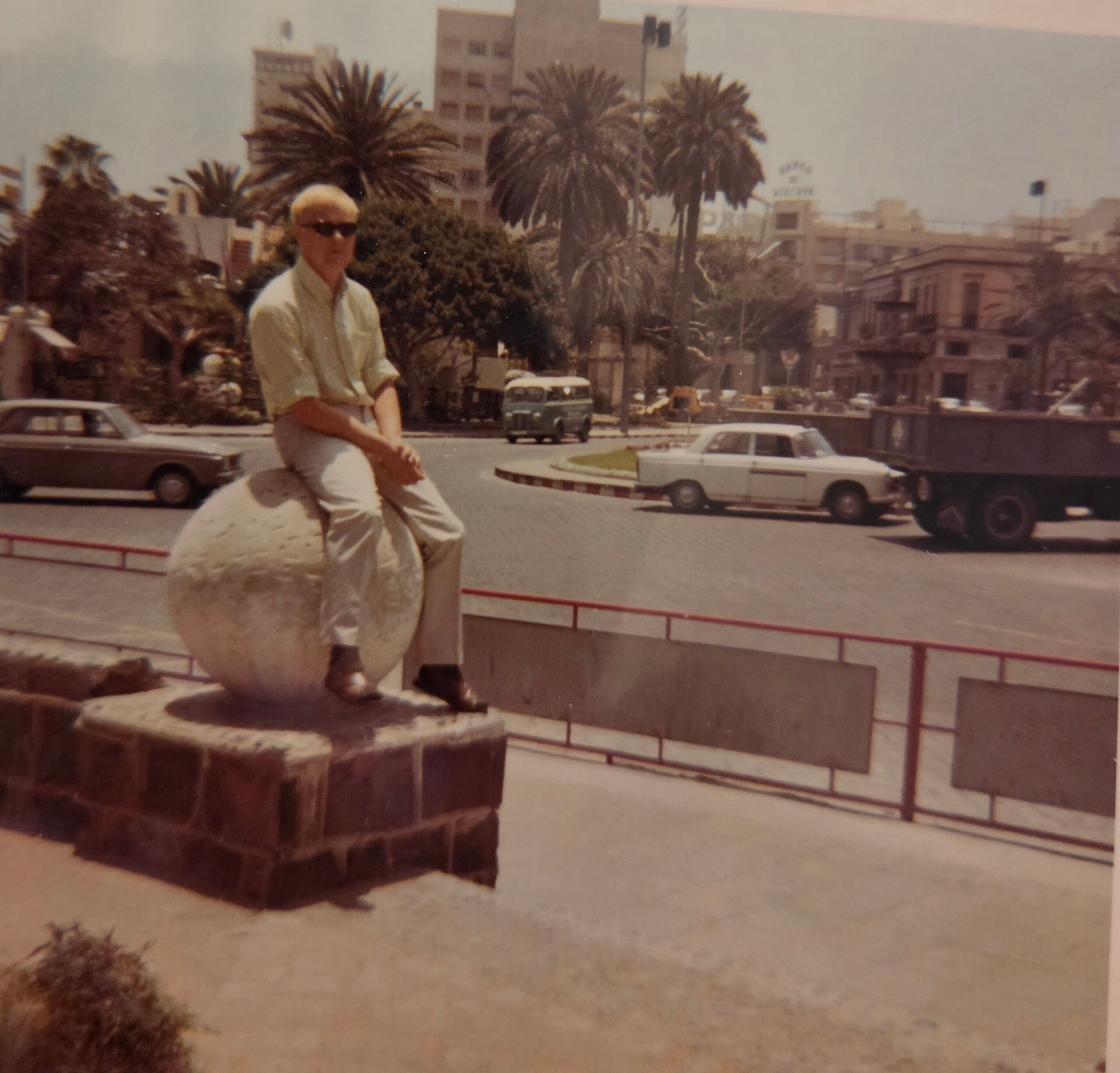 Older man in sunglasses sits on a stone sculpture in a sunlit city square with palm trees and traffic in the background.