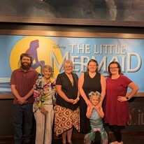 Six people posing together in front of a The Little Mermaid backdrop in a theater lobby, with a child in front raising arms.