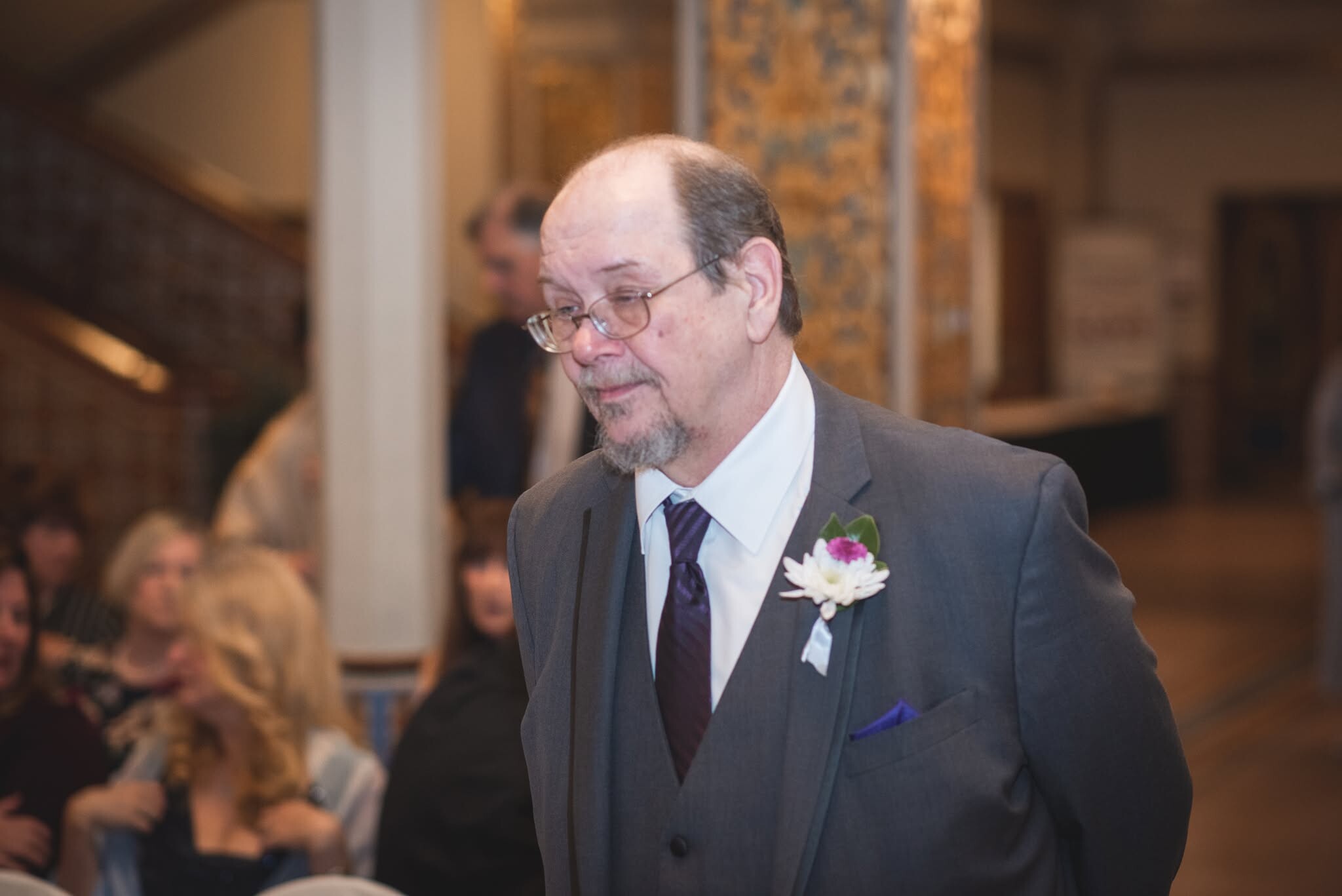 Man in a gray suit with purple tie and a flower boutonniere, looking down at a formal event with guests in the background.