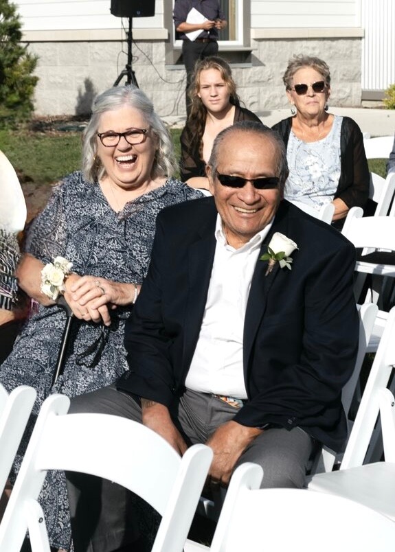 Smiling couple seated at a wedding ceremony, man in a suit with boutonniere and sunglasses, woman beside him with a corsage, outdoor chairs behind.