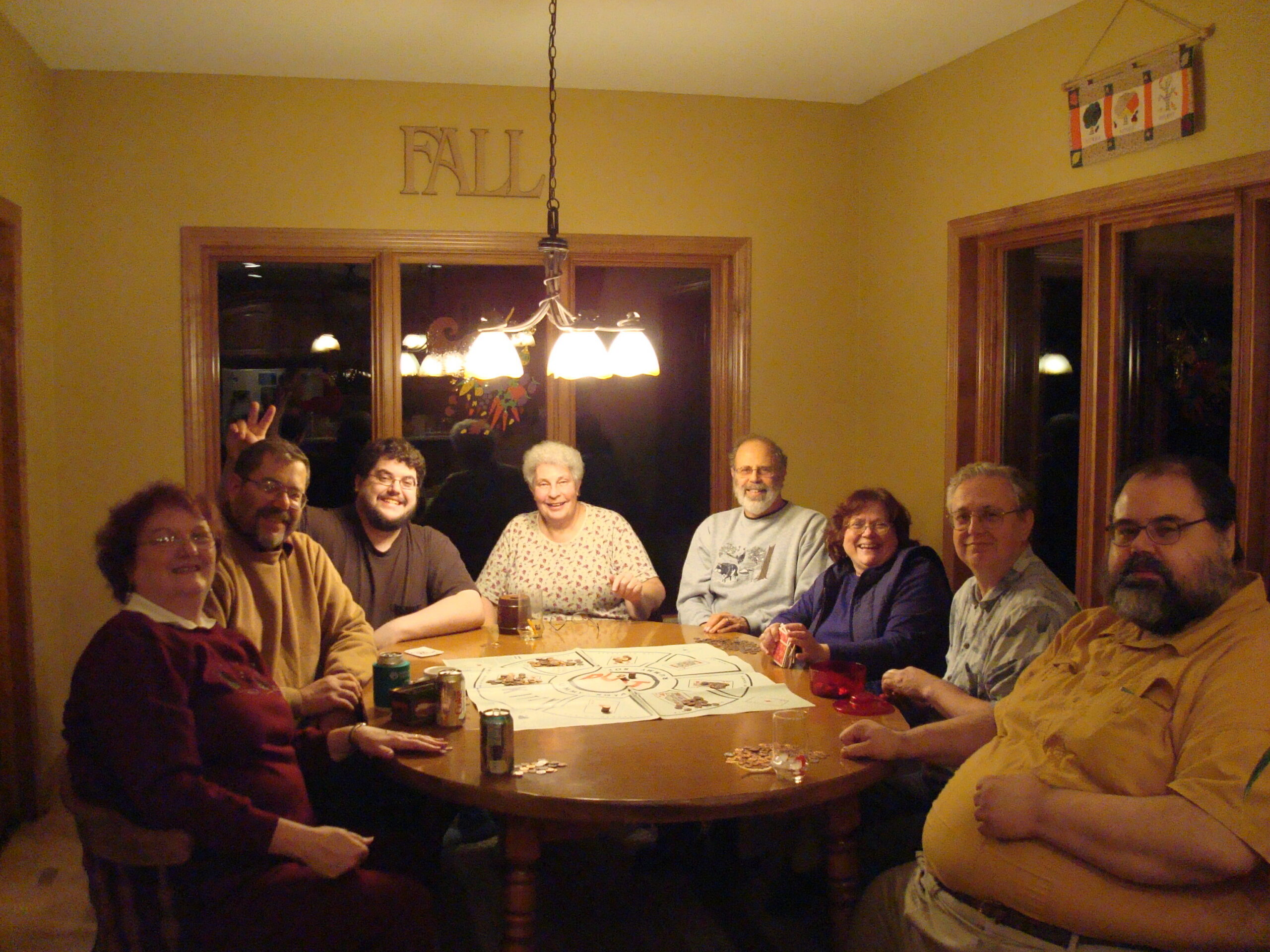 Group of adults seated around a dining table playing a board game, chips and drinks visible, warm kitchen lighting in background