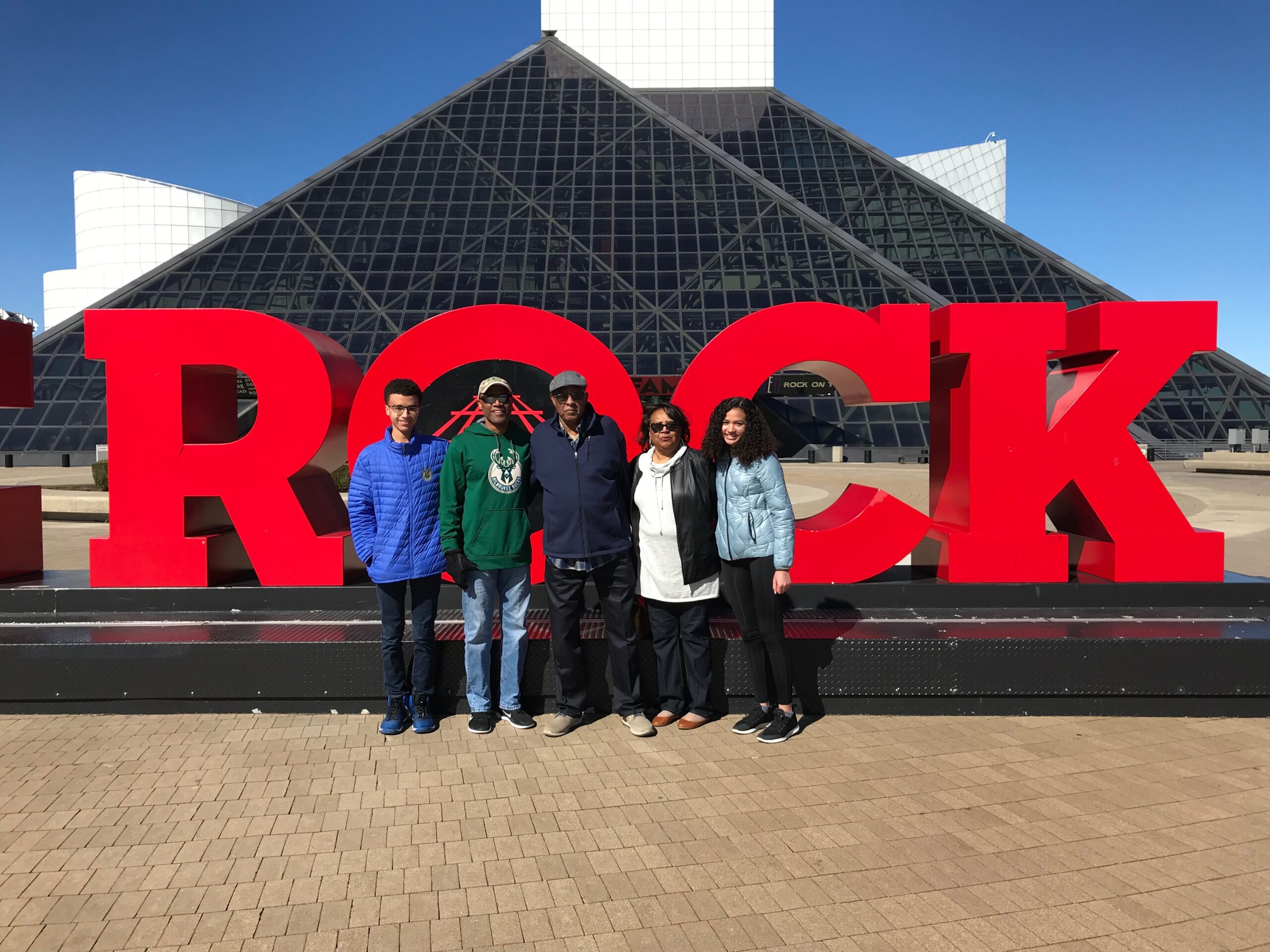 Five people stand together in front of large red letters spelling 'ROCK' at an outdoor museum entrance, with a glass pyramid building behind.