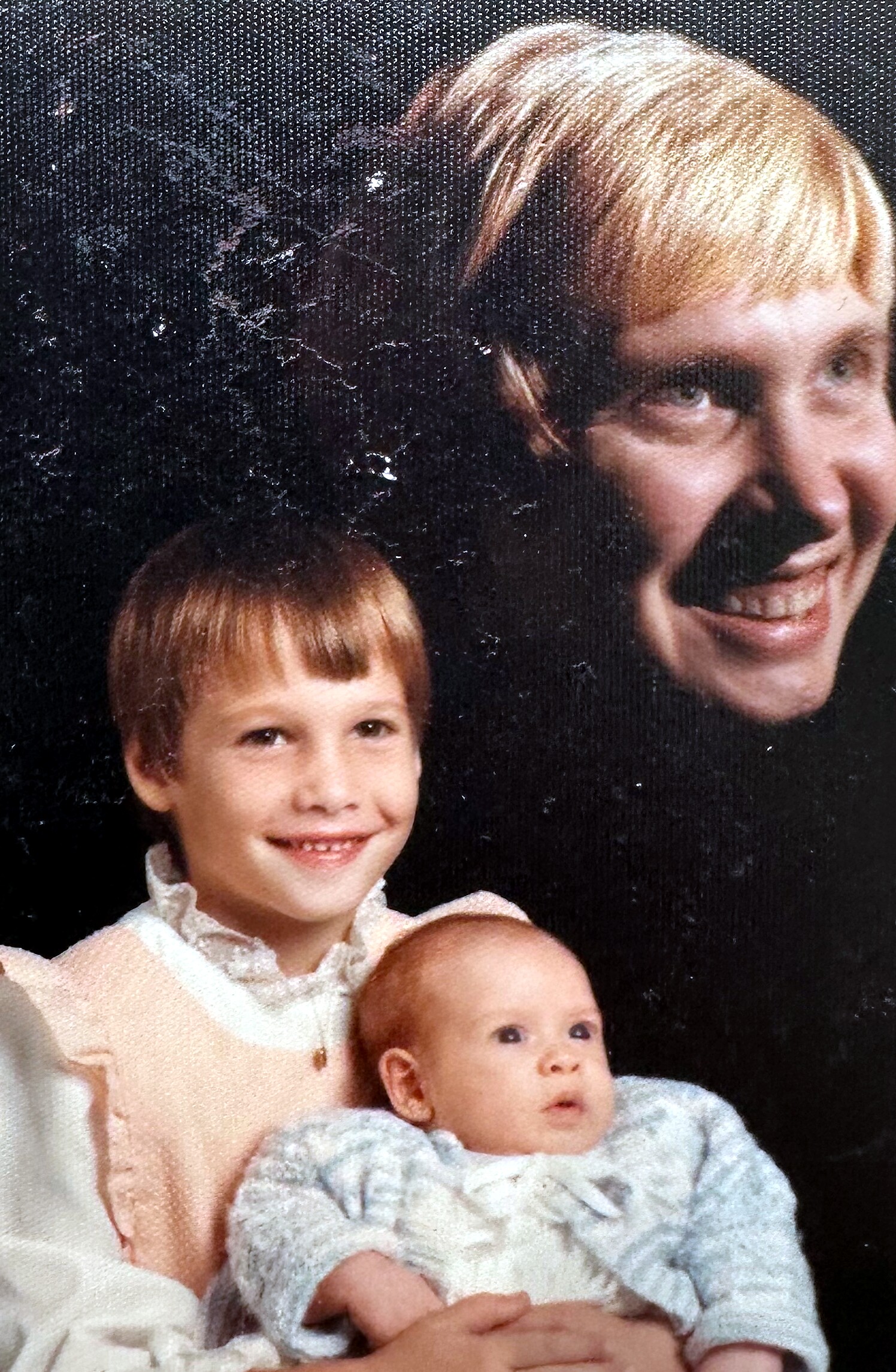 Smiling blonde woman holding a baby while a toddler stands beside her against a dark backdrop.