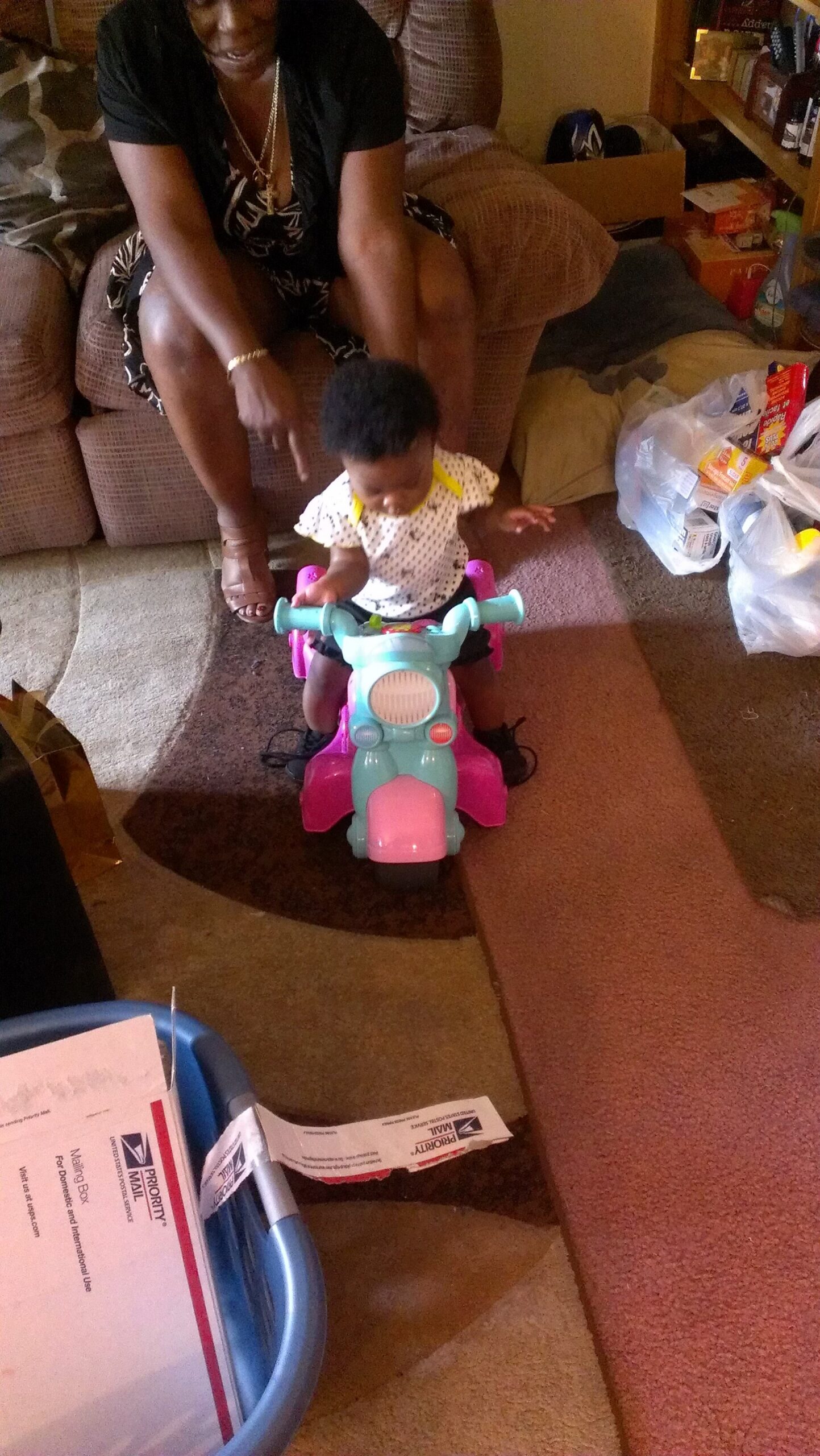 Child on a pastel ride-on toy with an adult woman guiding from behind in a cluttered living room.