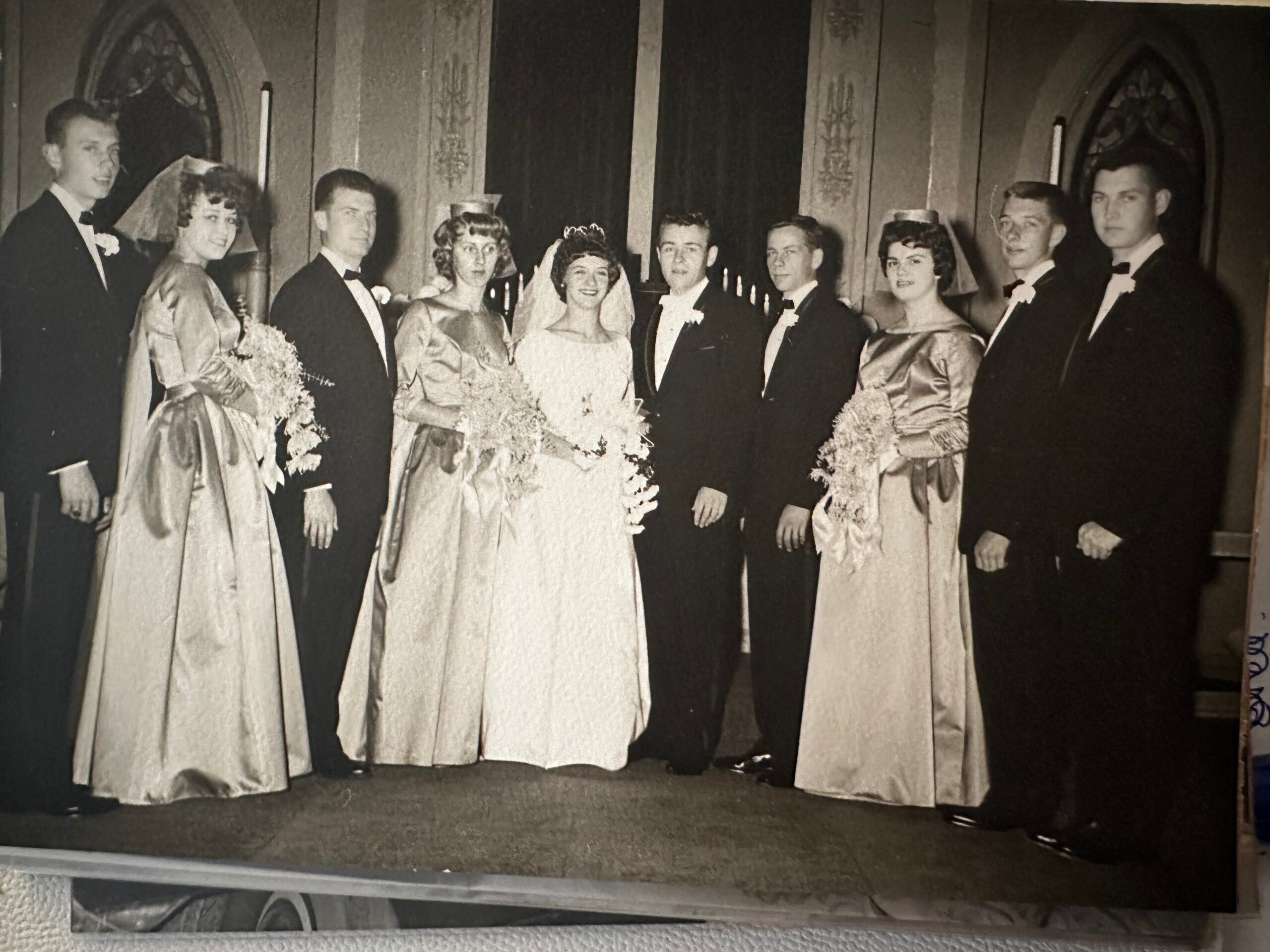 Group of bridesmaids and groomsmen posing in formal wedding attire inside a church hall.