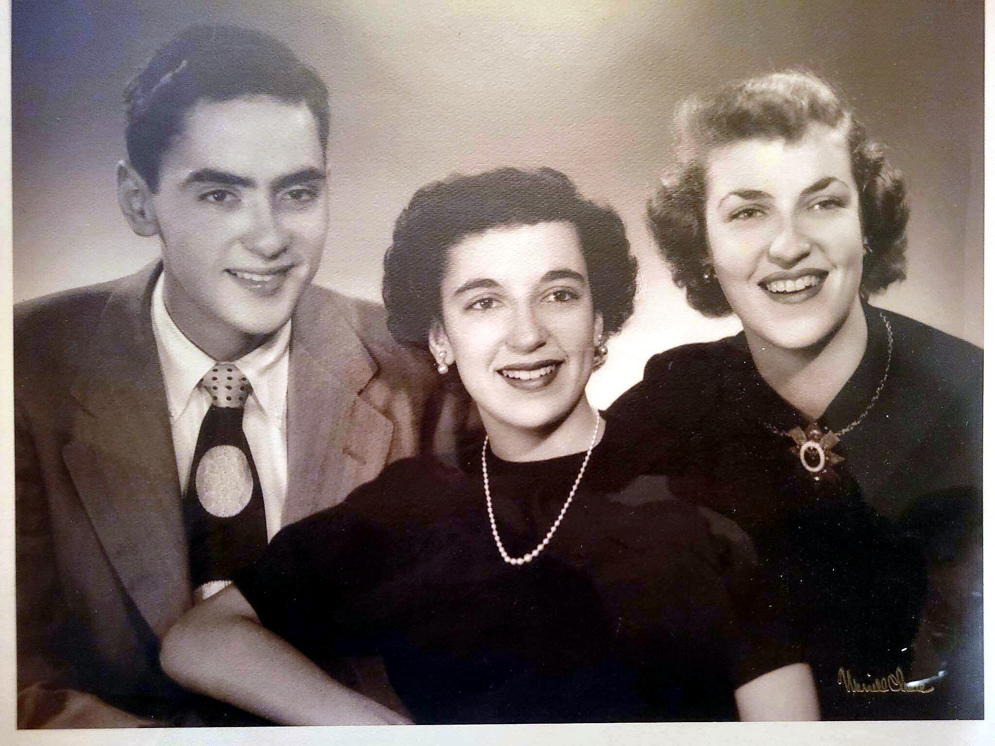 Three adults pose closely for a vintage studio portrait, smiling at the camera (man on left, two women center and right).