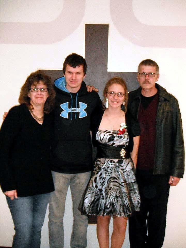 Group of four adults posing together indoors in front of a wall with a large cross in the background.