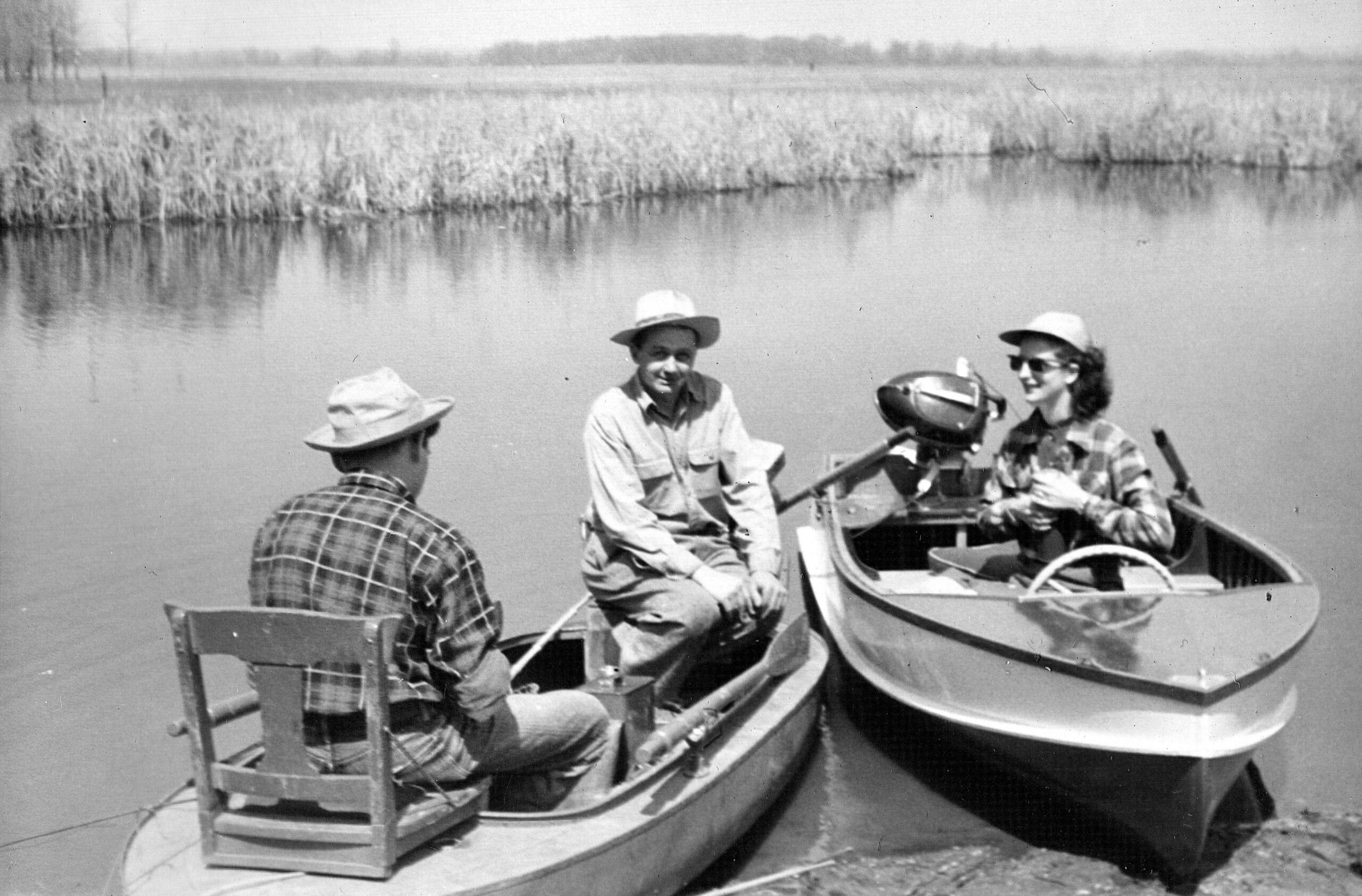 Three people in a small rowboat on a calm lake; a man in a hat sits near the middle while another woman wears sunglasses and a hat at the stern-like area, with a paddler in a plaid shirt nearby.
