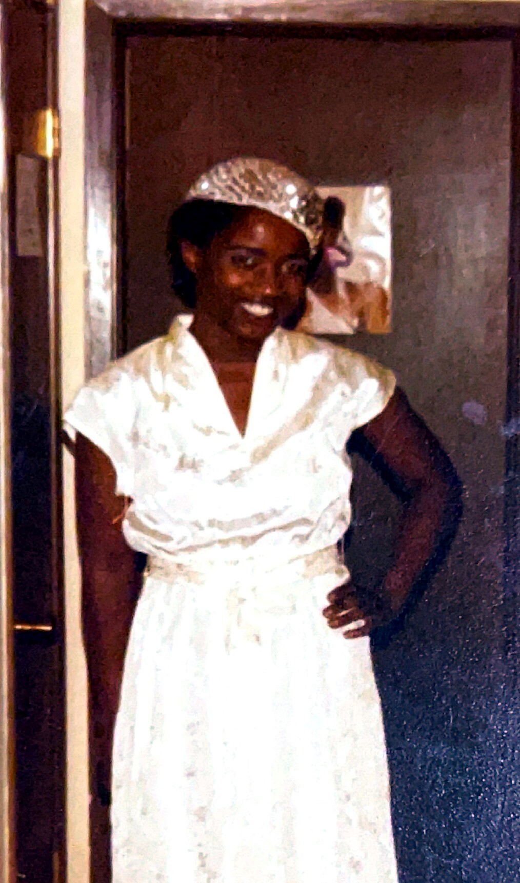 Smiling woman in a white dress and silver head wrap posing with her hand on her hip in a doorway area.
