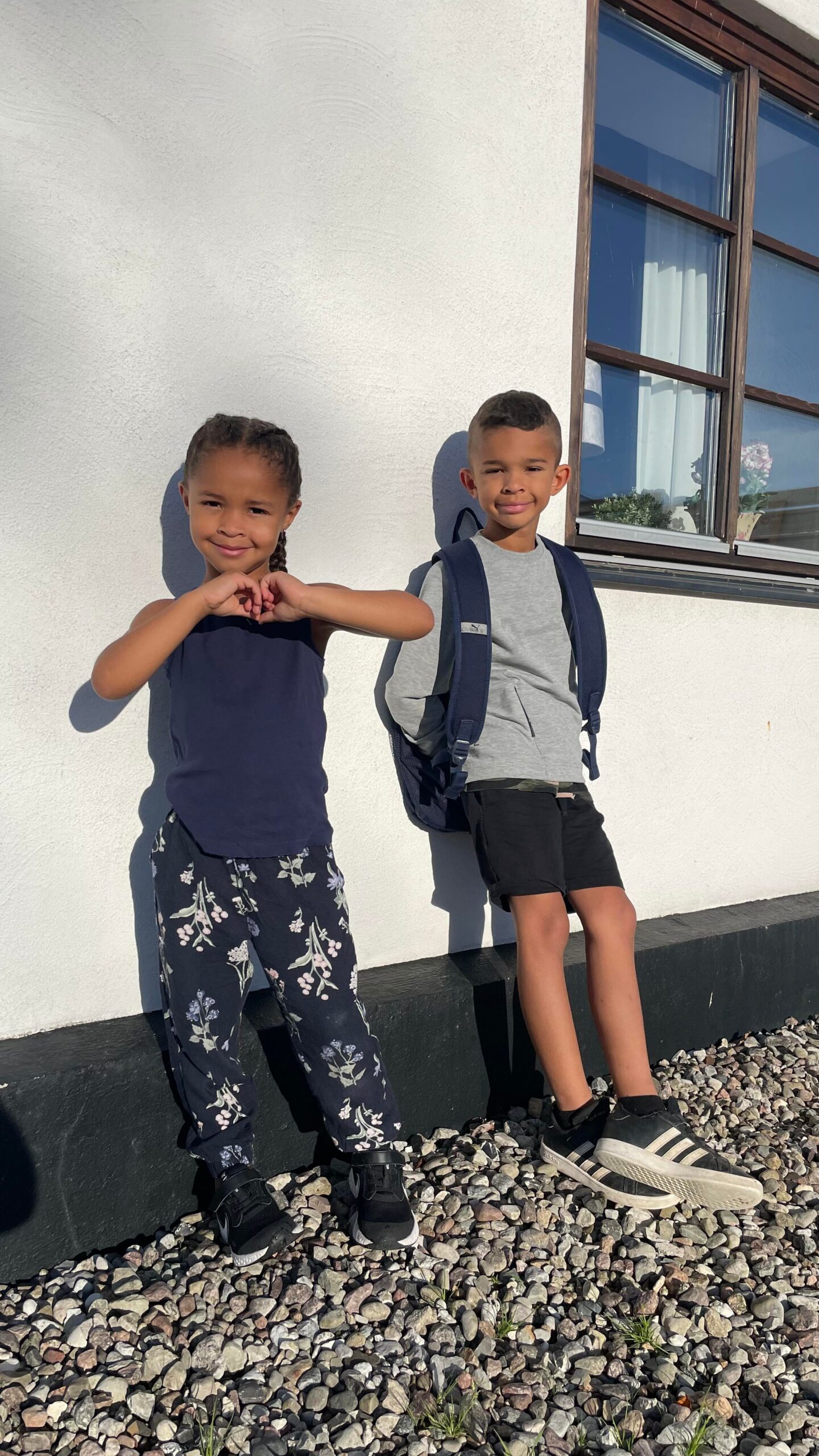 Two kids stand against a white wall on a rocky ground; girl on the left forms a heart with her hands, boy on the right wears a backpack.