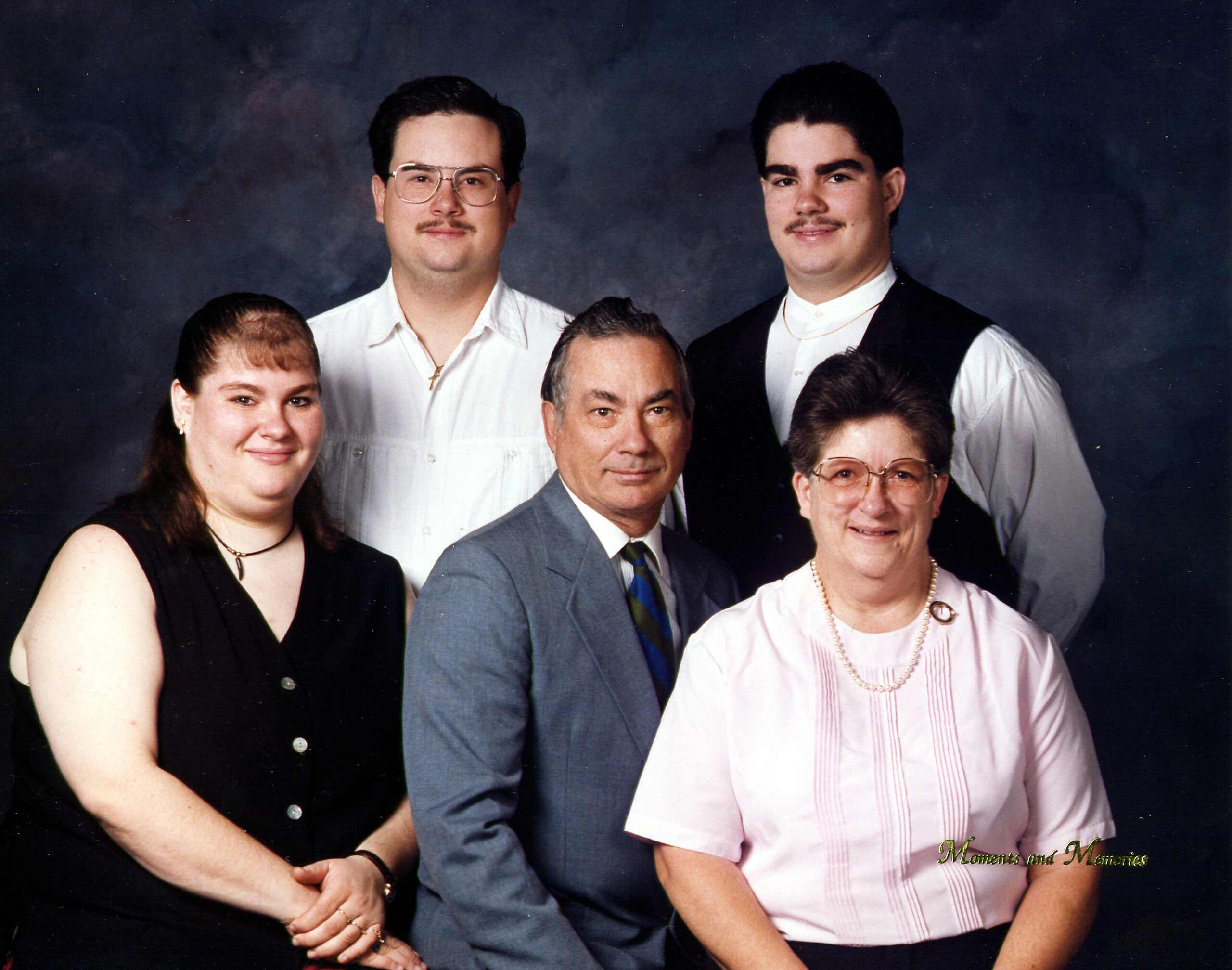 Group family portrait in a studio: five relatives in formal attire posing against a dark backdrop, with two men standing behind and three seated in front.