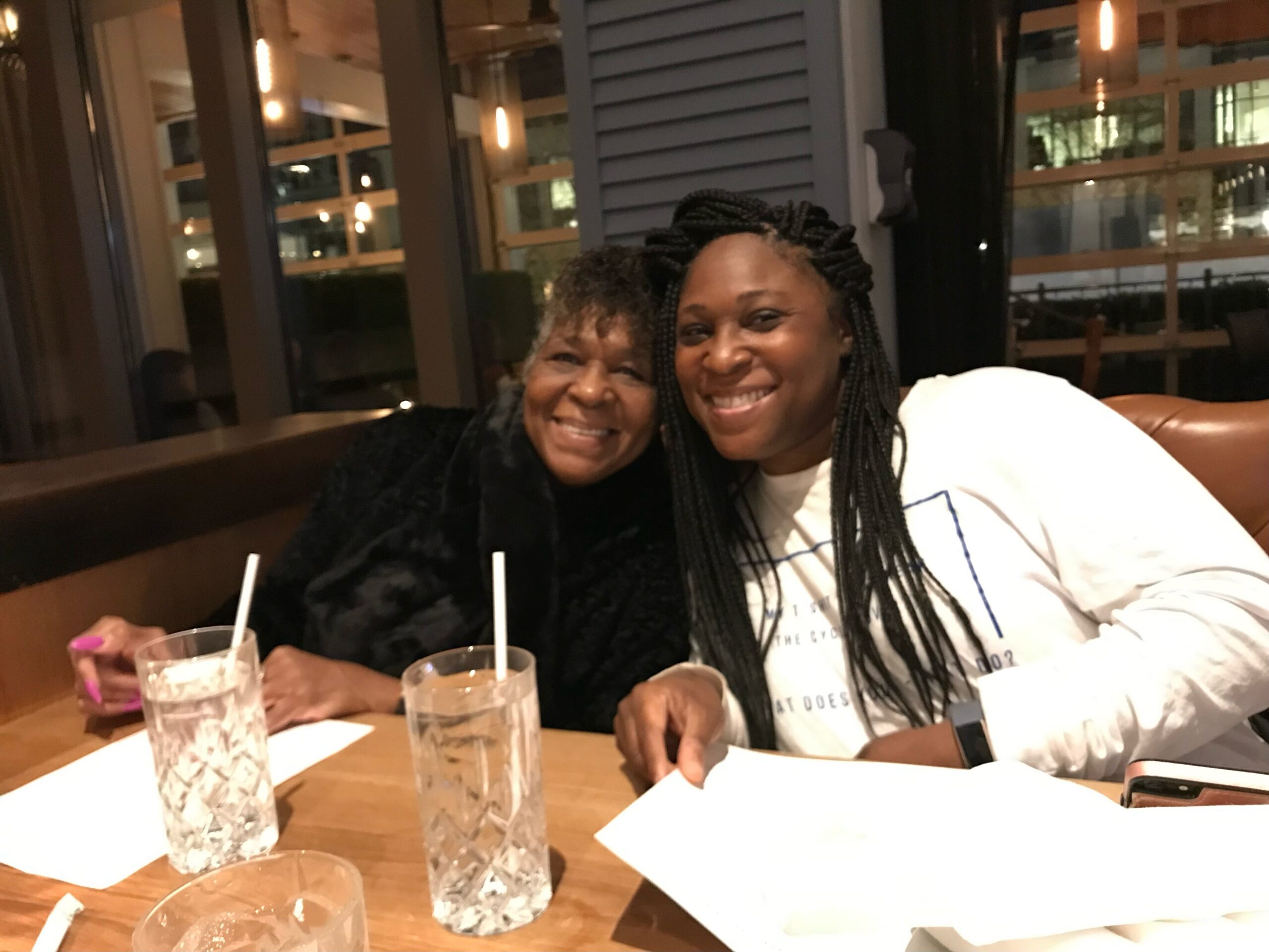 Two women smiling at a restaurant table with drinks and papers in front of them at night (indoor setting).