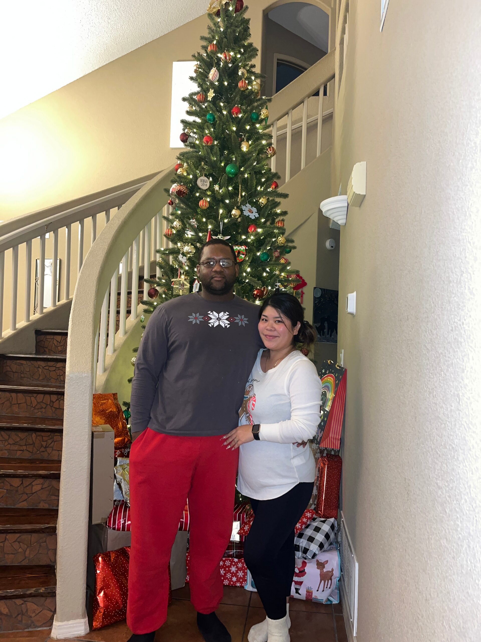 A couple posing indoors next to a tall decorated Christmas tree with wrapped gifts beneath it and stairs in the background.