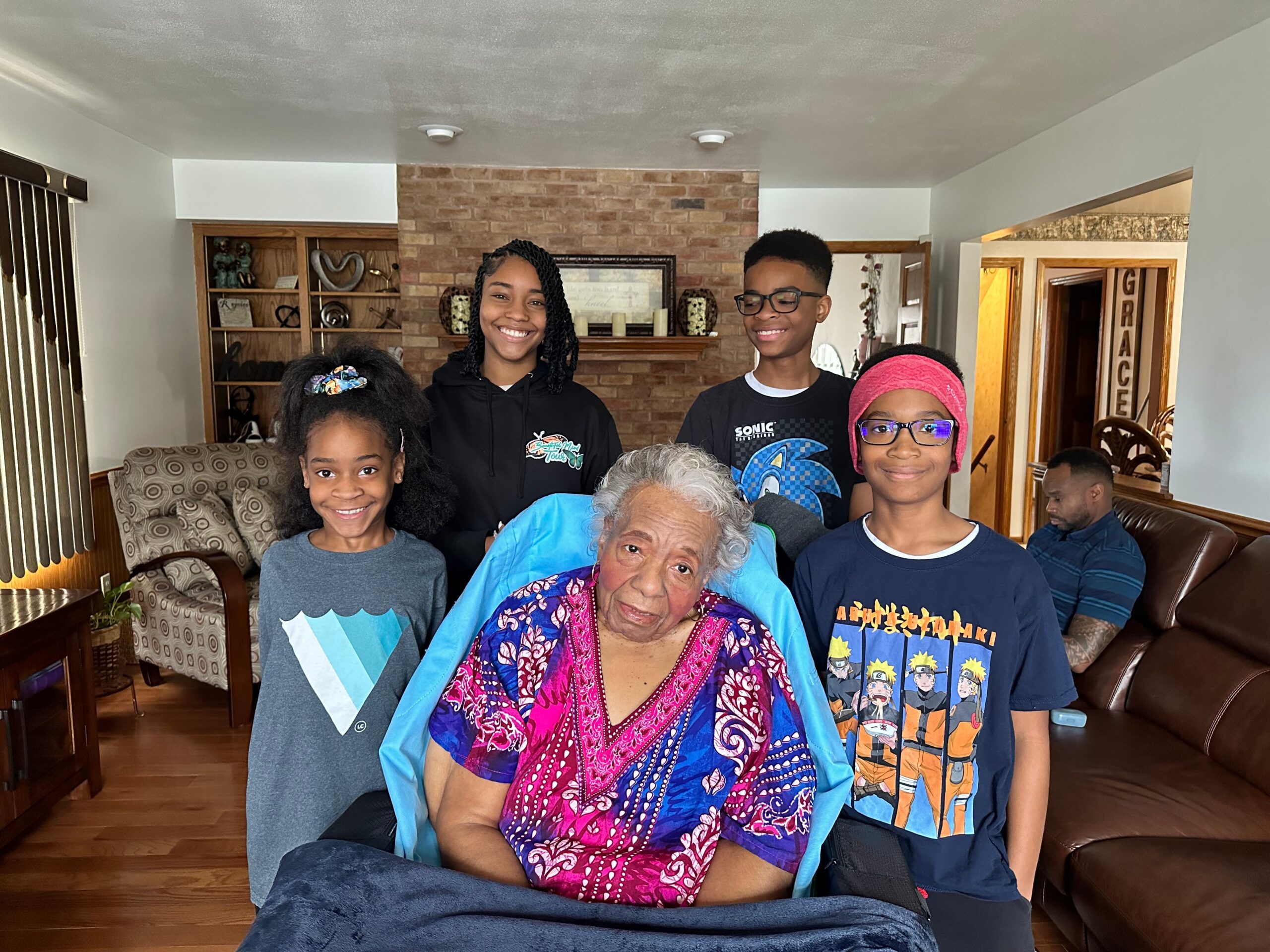 Multi-generational family posing indoors in a living room with a brick fireplace behind them, smiling at the camera.