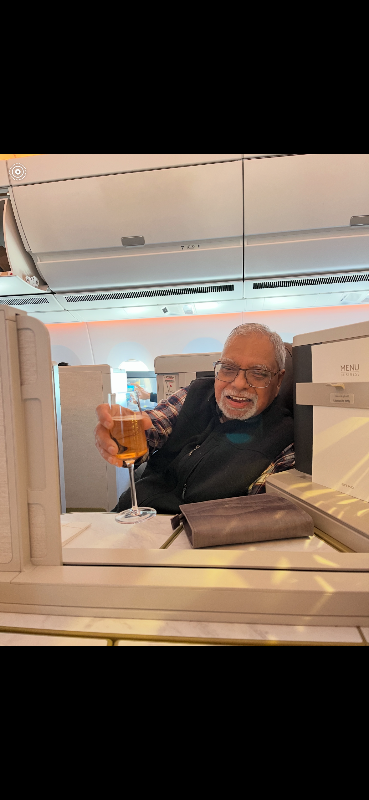 Older man with glasses and a white beard sits in a modern airplane cabin, holding up a glass of amber beverage and smiling.