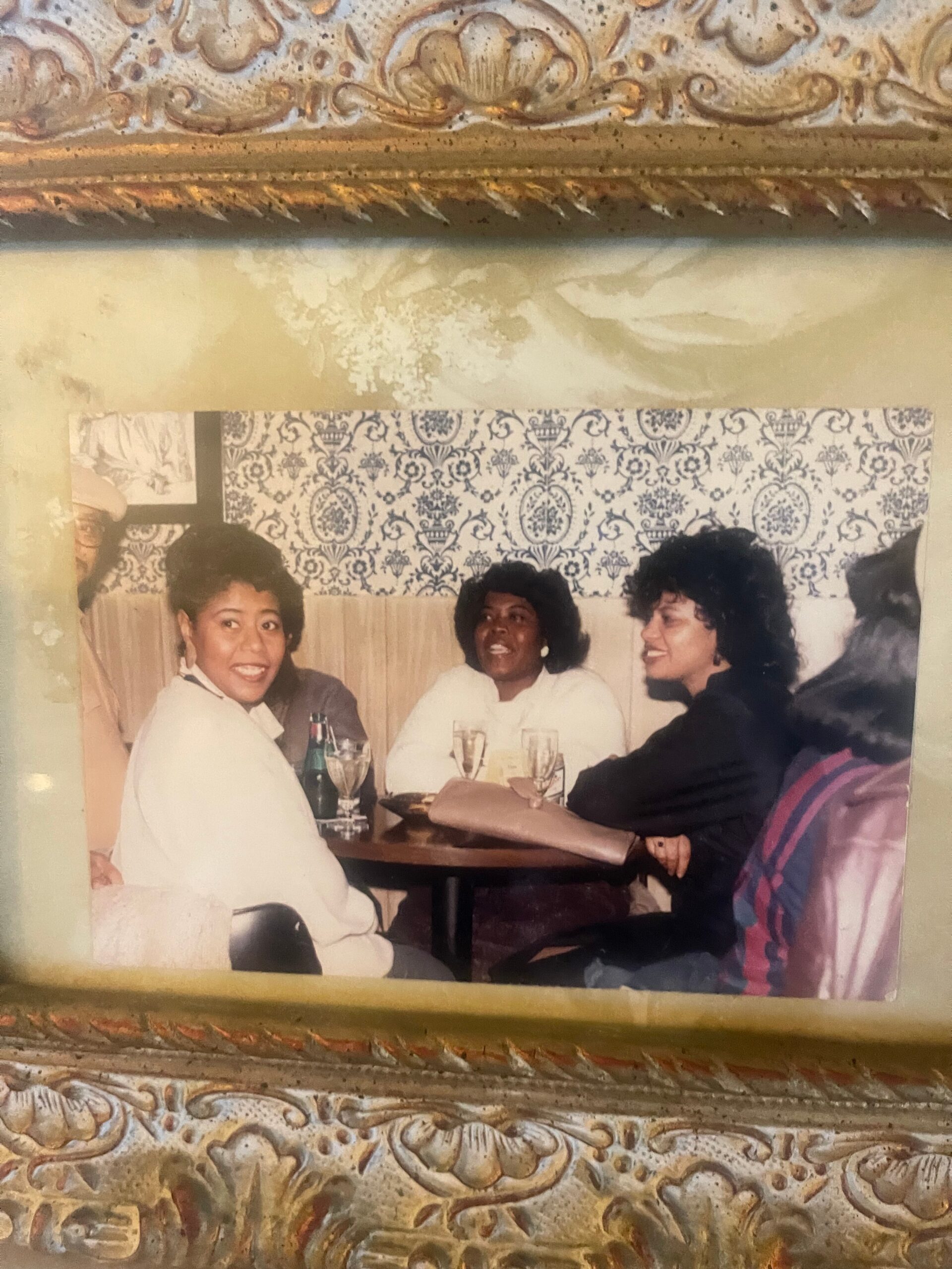 Three women sit at a round table in a restaurant, smiling and chatting, with drinks visible on the table and a patterned wall behind them.