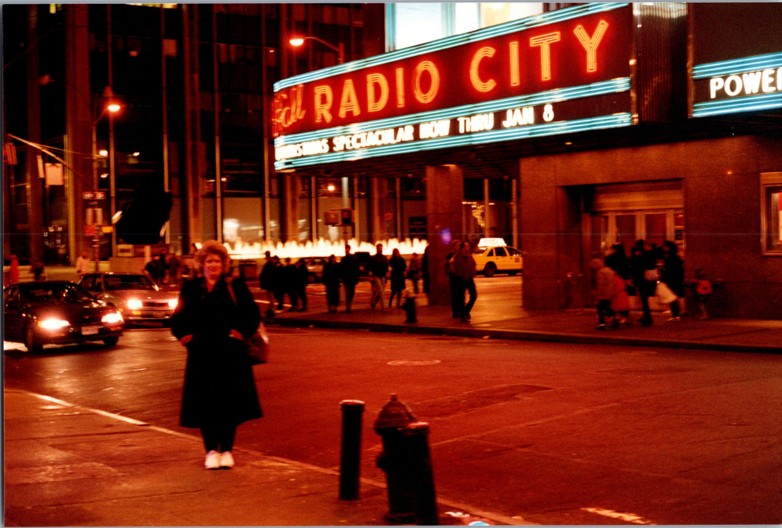Night scene outside Radio City with a neon marquee; pedestrians, cars, and a lit fountain on a wet street at night.