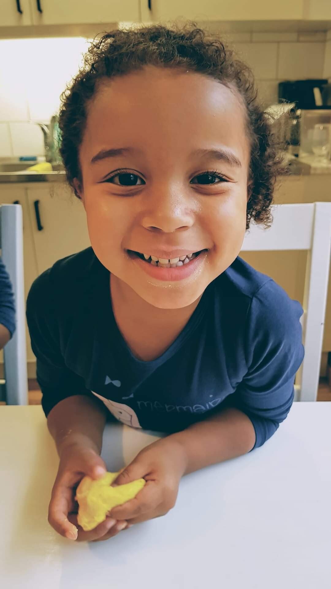 Smiling child with curly hair sitting at a table, shaping yellow playdough in a kitchen.