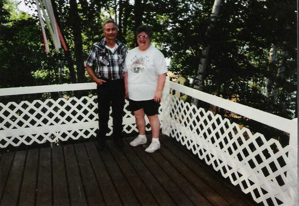 Two adults pose on a wooden deck with a white lattice railing, smiling, with trees in the background.