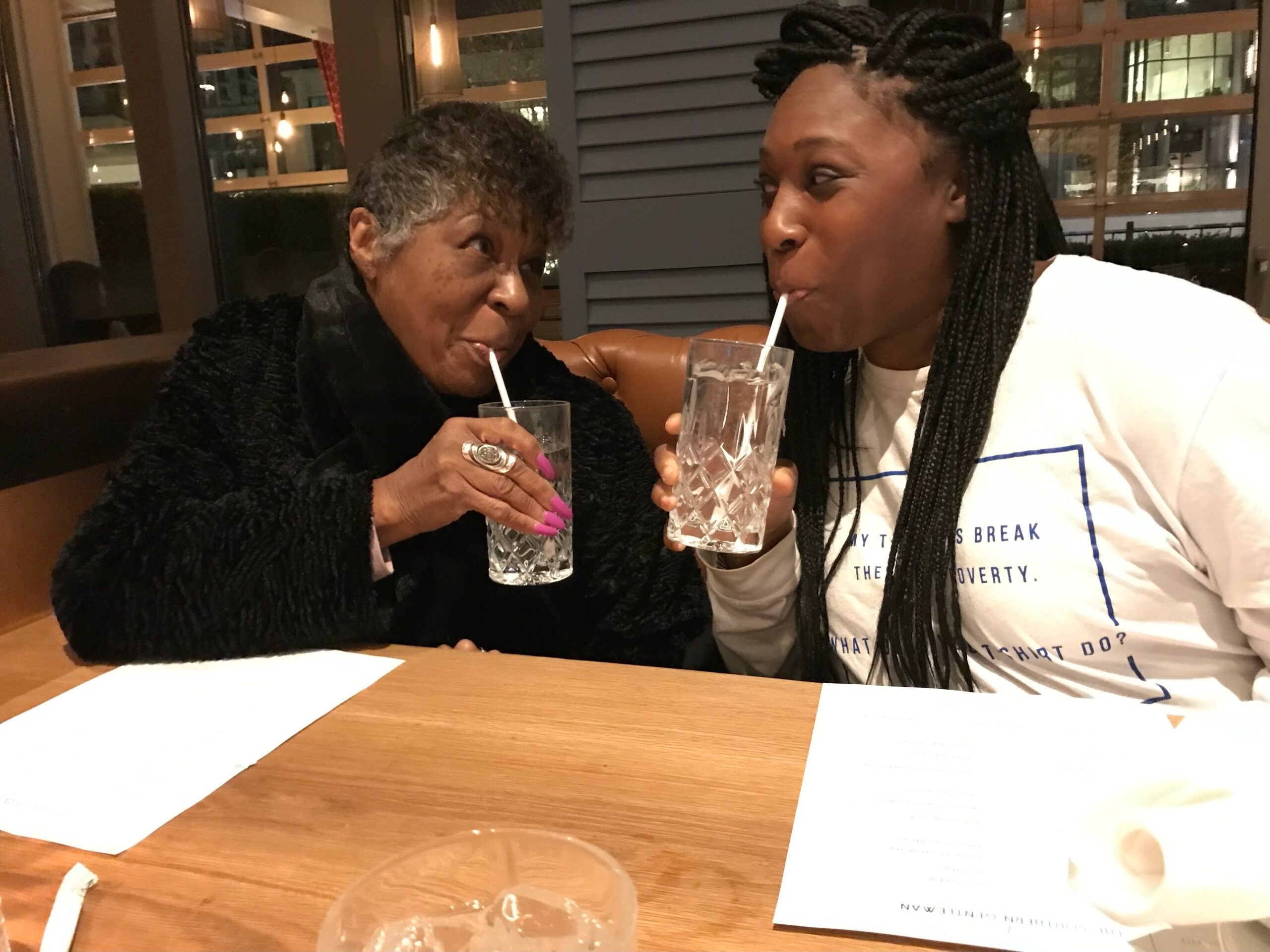 Two women sit at a restaurant table, each sipping from a tall glass with a straw and smiling at each other on a night out.