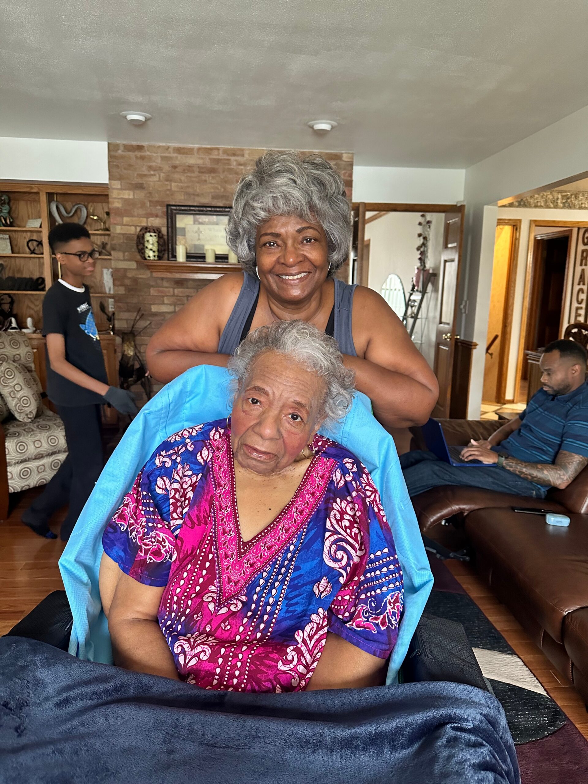 Smiling elderly woman in a blue shawl seated, with a gray‑haired caregiver standing behind her in a cozy living room.