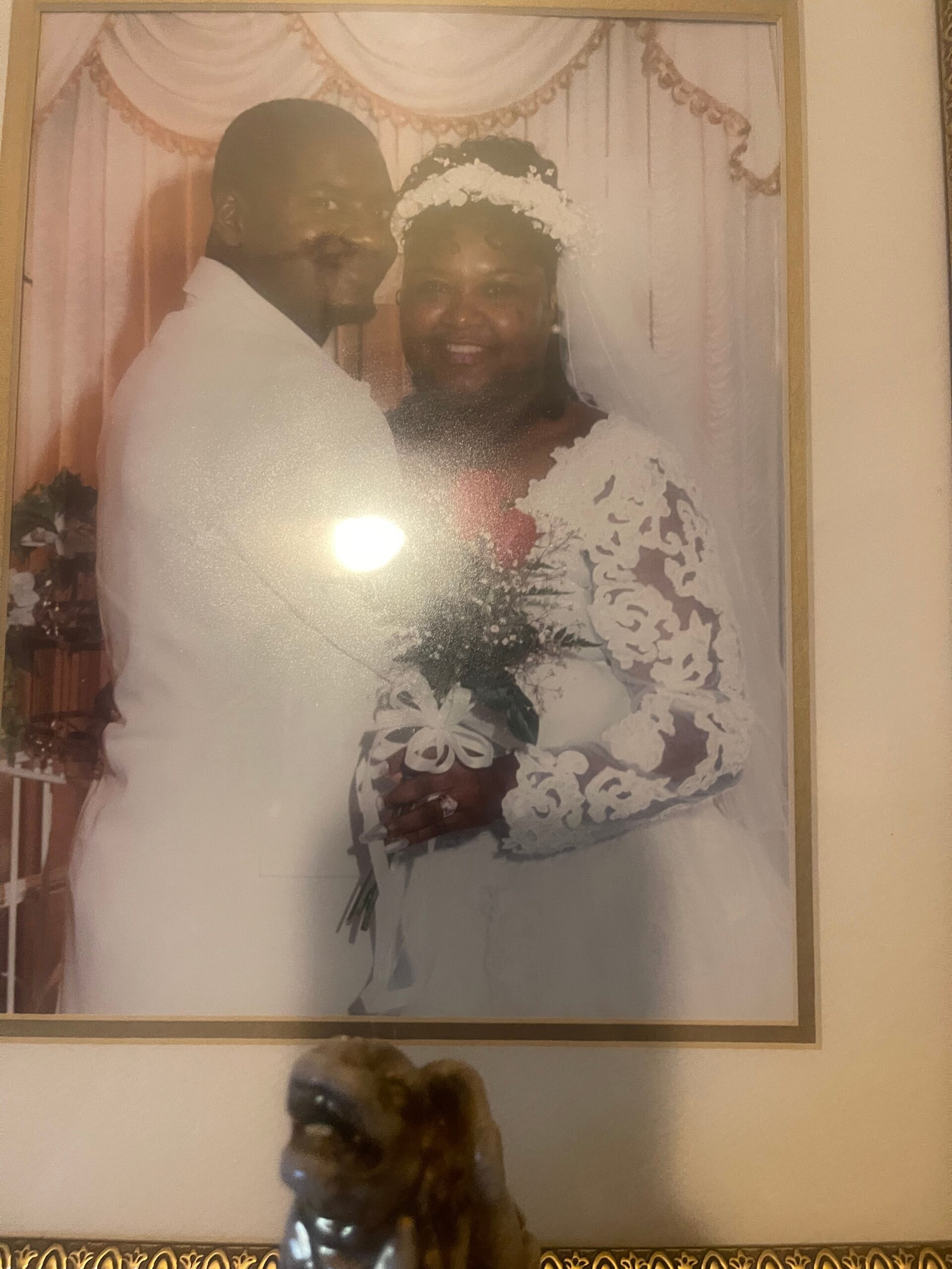 Framed wedding portrait of a smiling couple: bride in lace gown and veil with a bouquet, groom in a white suit beside her in front of a pink backdrop.